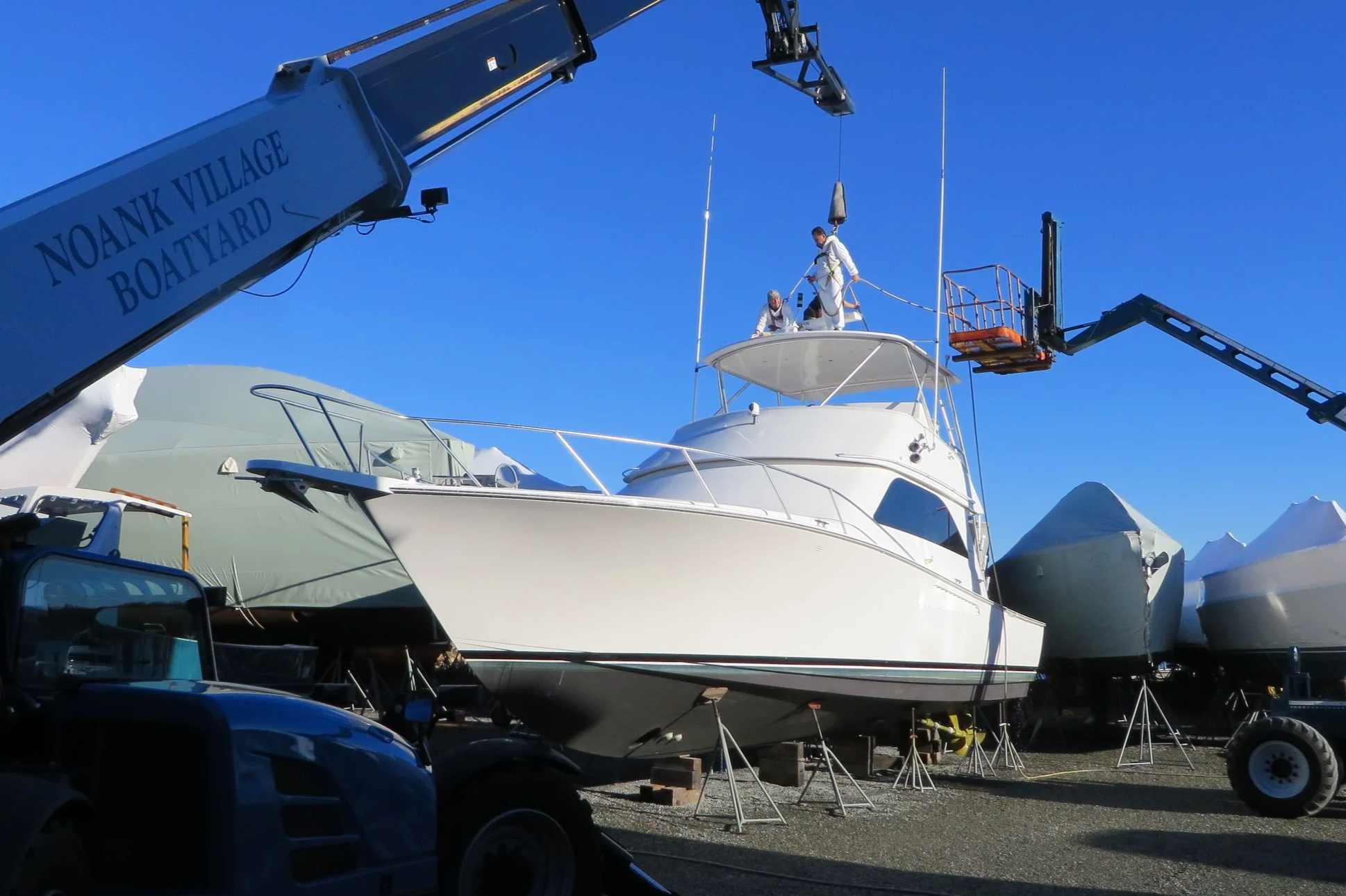 People working on a boat with a crane at a boatyard, with other boats covered and stored nearby, under a clear blue sky.