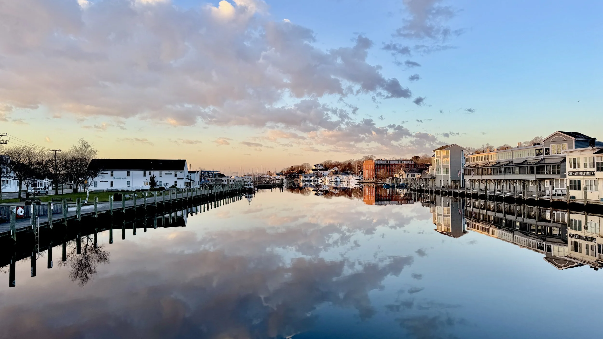 A peaceful harbor scene at sunset with calm water reflecting the sky, boats, and buildings along the waterfront, some with signs like 'Steamboat Inc.'