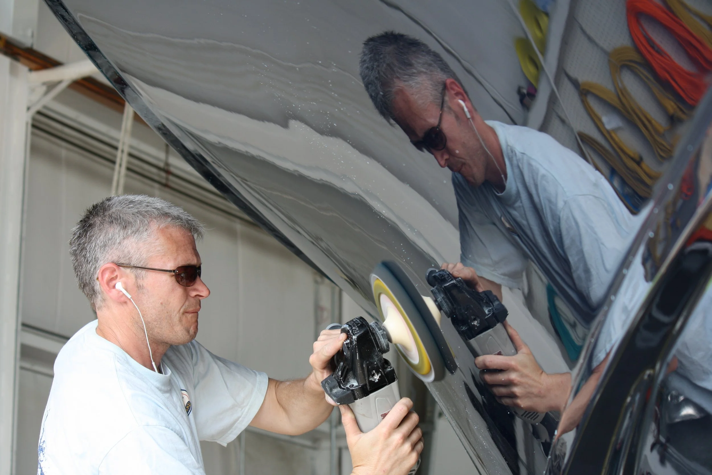 Two men working on polishing or buffing the exterior of a black car inside a garage. They are using handheld power tools, and one man is wearing sunglasses and listening to music with earphones.