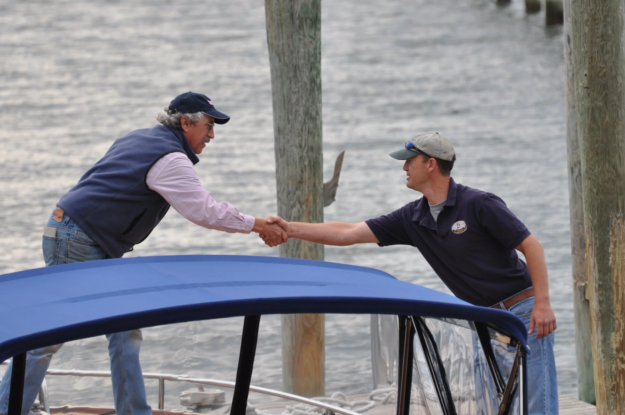 Two men shaking hands on a dock by the water, with wooden pilings in the background, one man wearing a navy vest and cap, and the other in a dark polo shirt and cap.