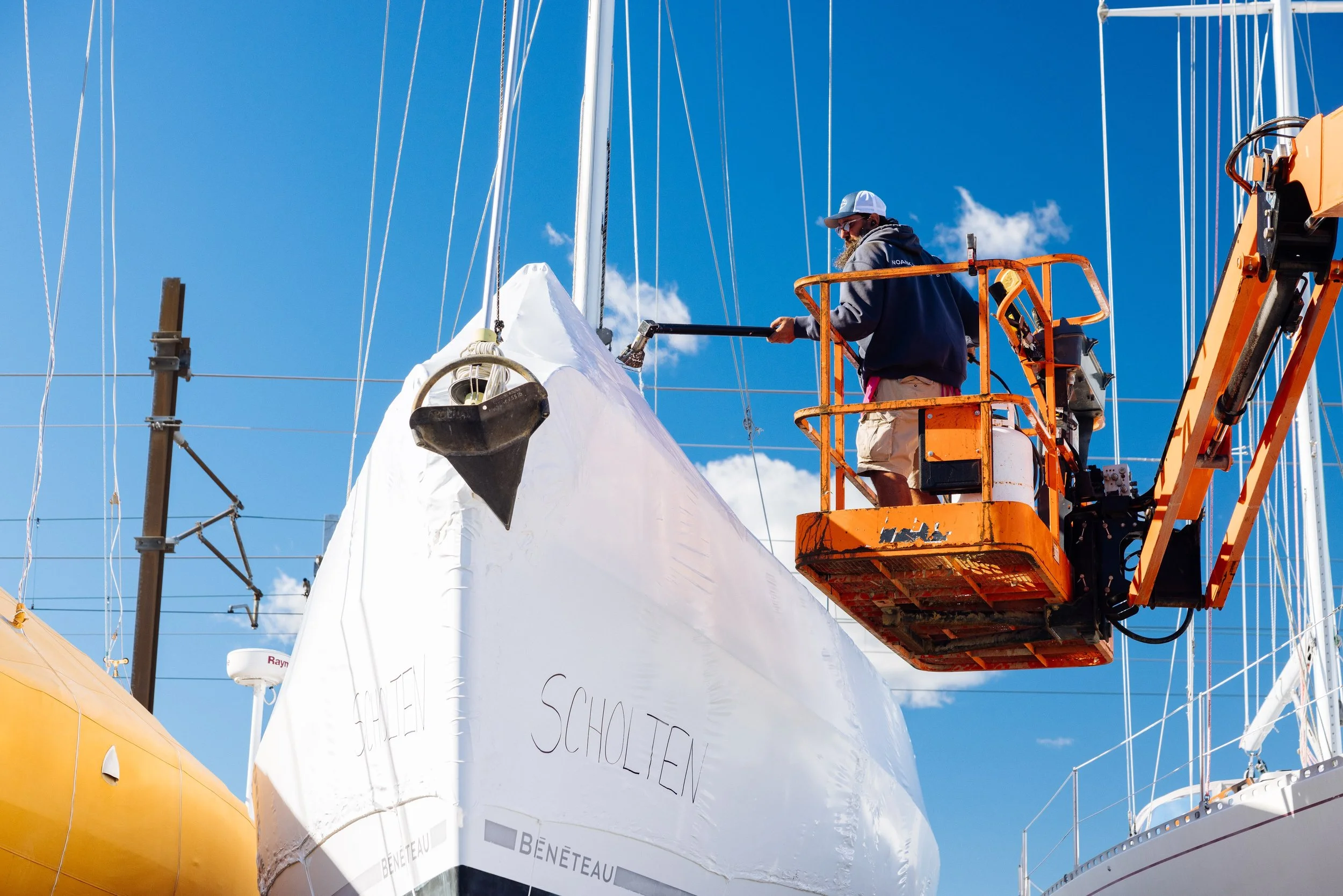 A man in a gray hoodie, beige shorts, and a white cap standing on an orange aerial lift working on a large sailboat's bow, against blue sky with a few clouds.