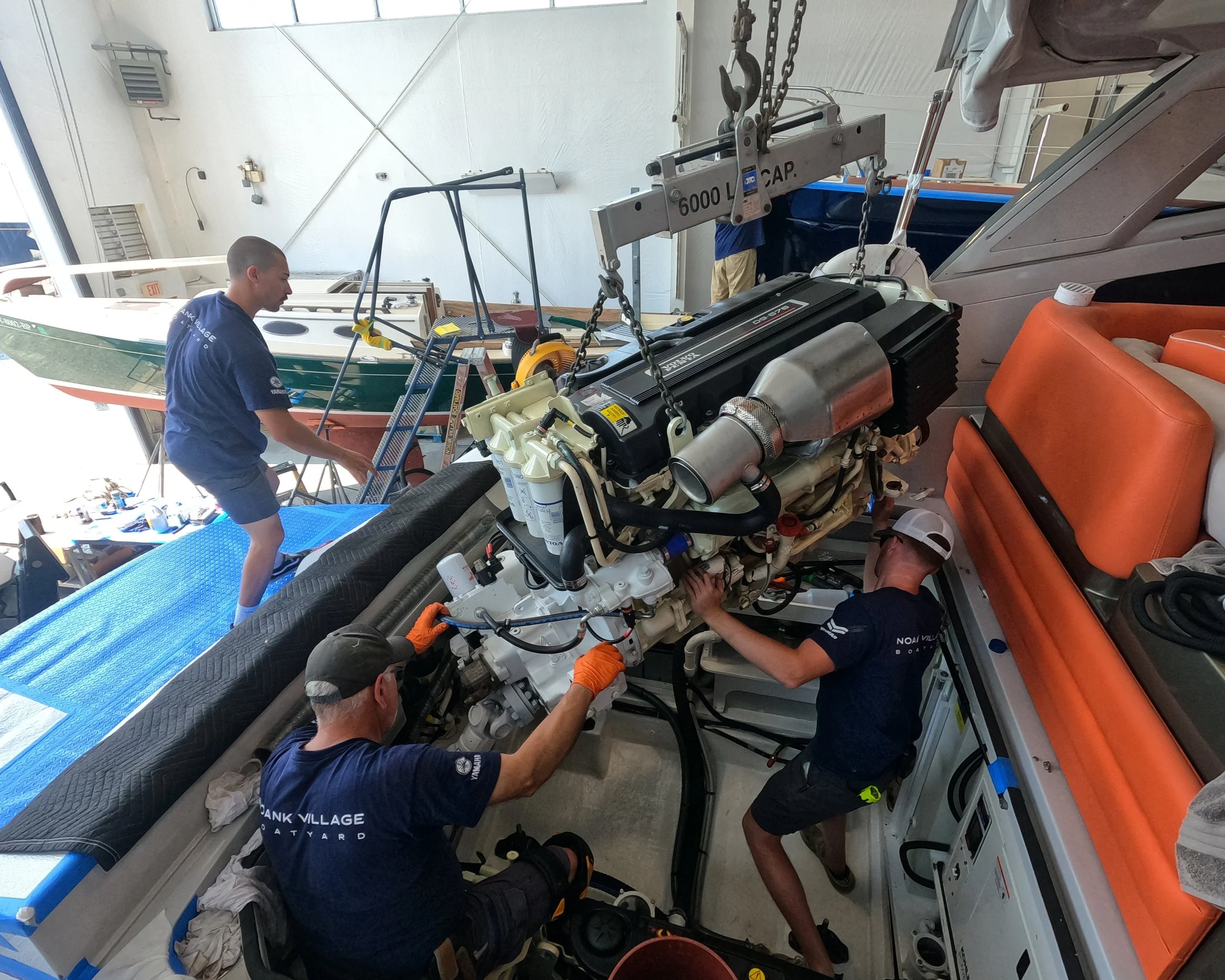 Multiple technicians working on a boat engine inside a workshop. The engine is large and complicated, with several wires and parts visible. Some workers are using tools to work on it, and there is a blue platform and an orange chair nearby.