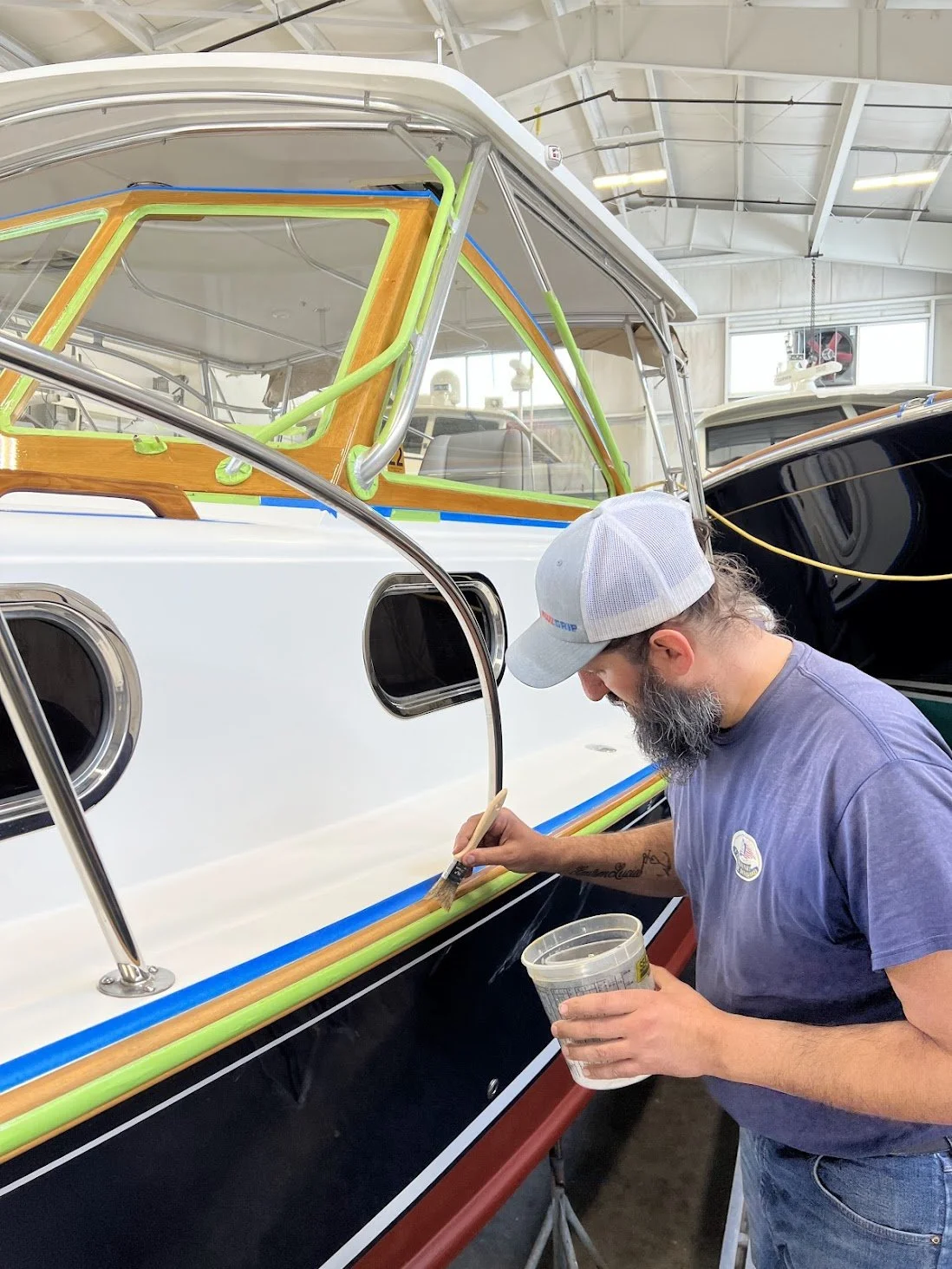 Man painting the side of a boat inside a workshop, wearing a gray cap and a blue T-shirt.