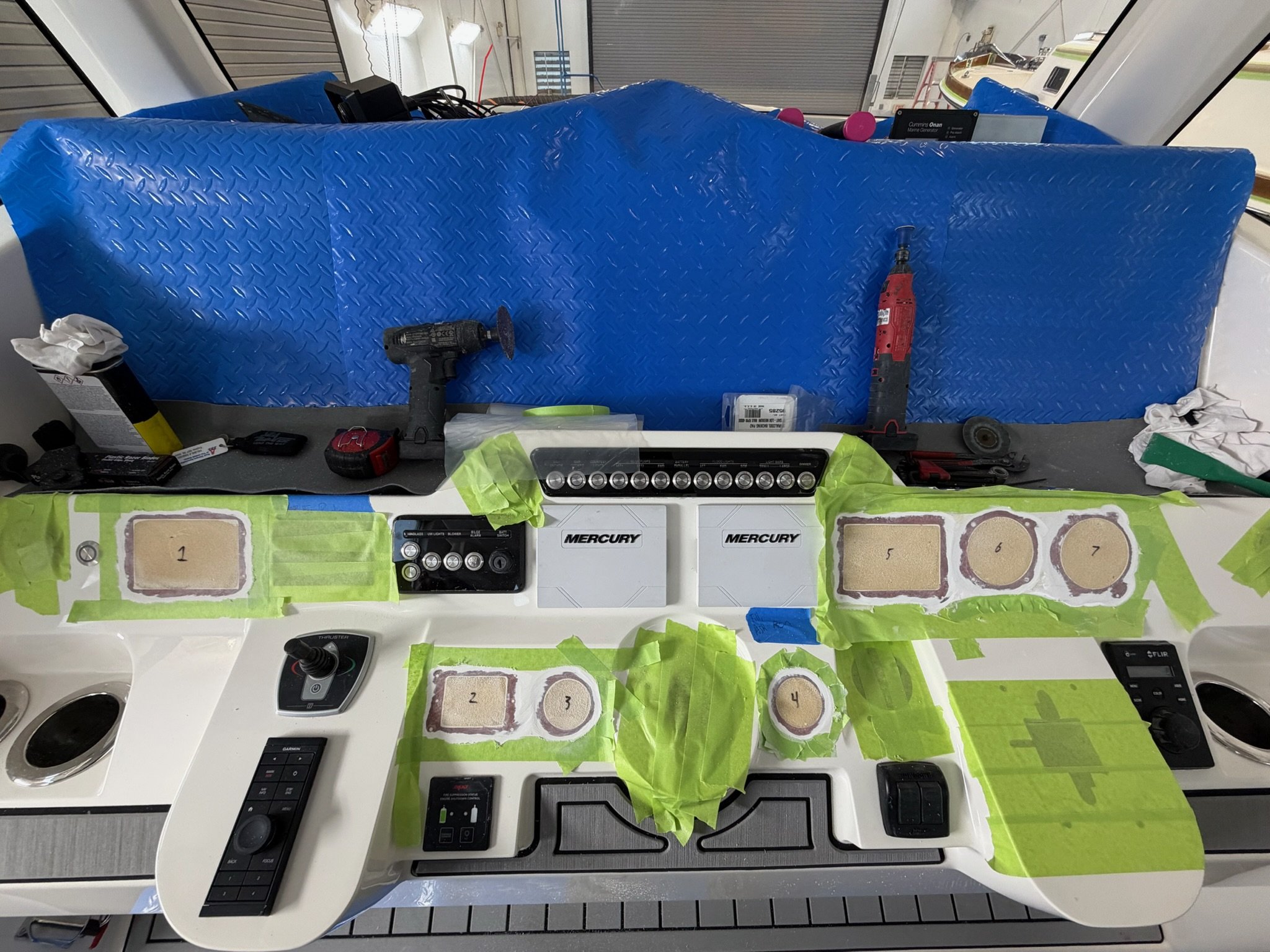 A workspace with tools and materials for boat or marine work, including drills, screwdrivers, an LED light bar, and numbered samples of a material, with a blue protective cover and a background of a boat or yacht interior.