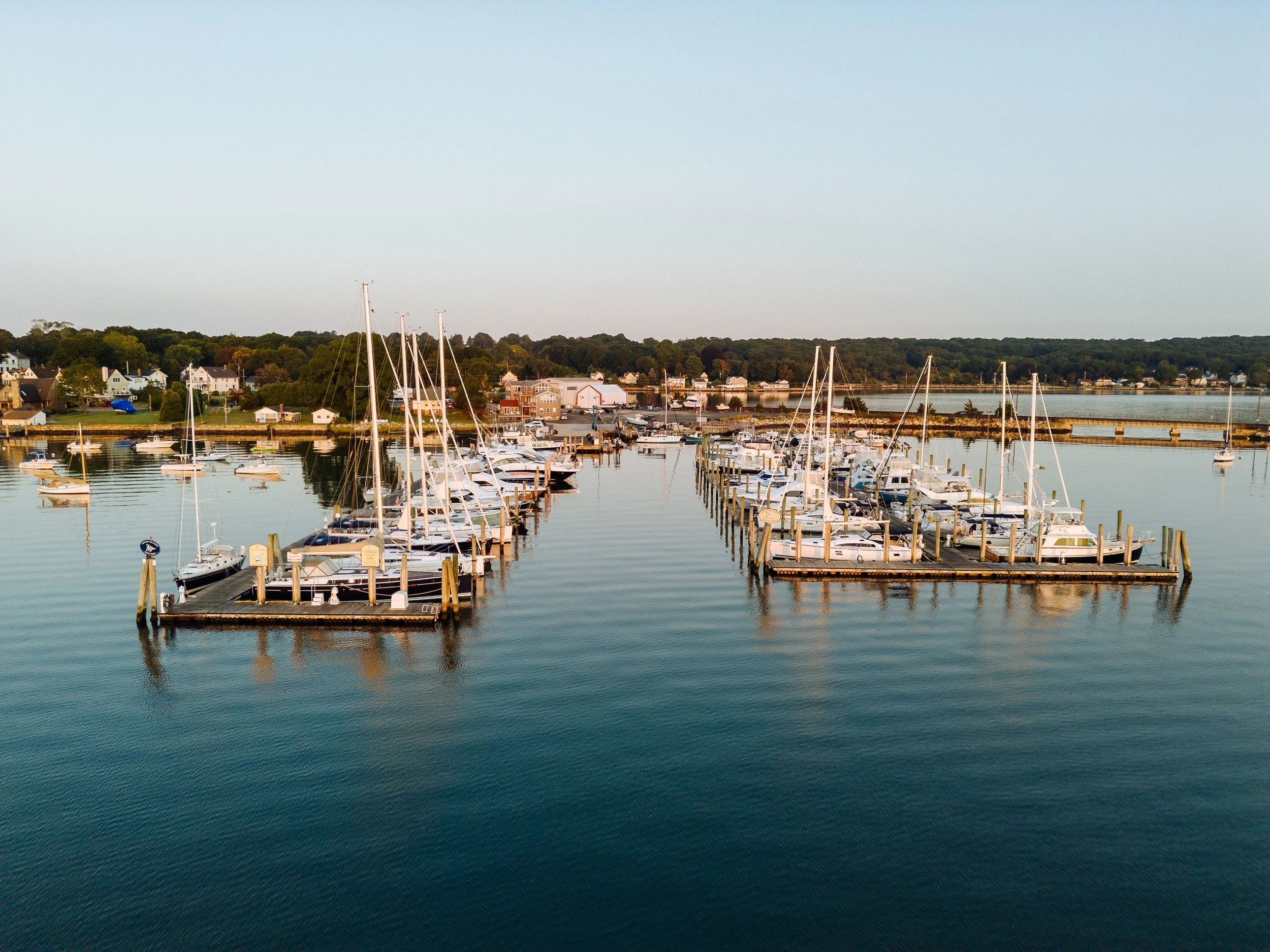 Marina with sailboats and yachts docked on calm water, houses along the shoreline, forested hillside in the background, clear sky.