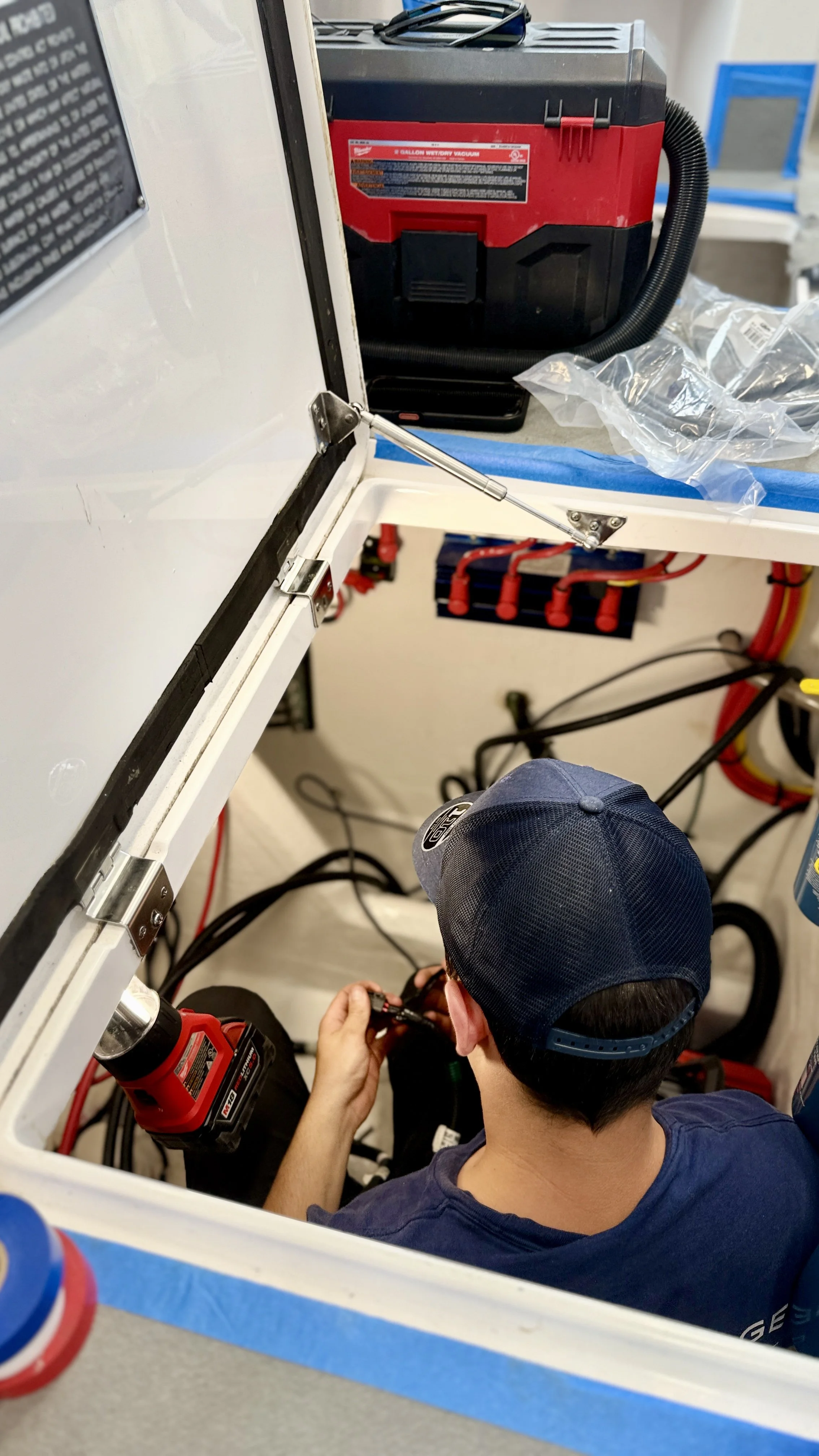 A person wearing a navy blue baseball cap working on electrical wiring inside a white enclosed space with tools and equipment around.