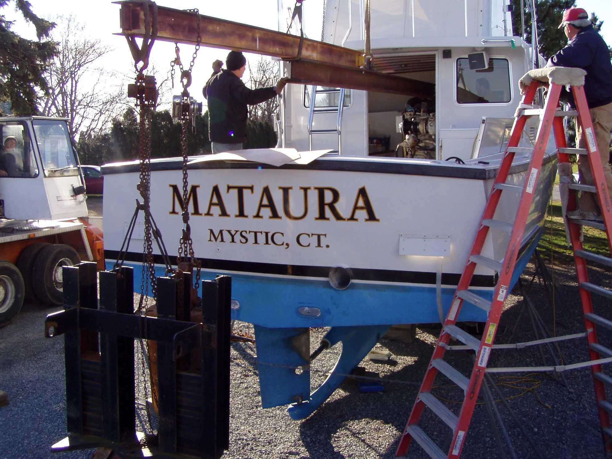 People working on a boat named 'Mataura' in a boatyard, with a crane and ladder nearby, preparing for maintenance or repair work.