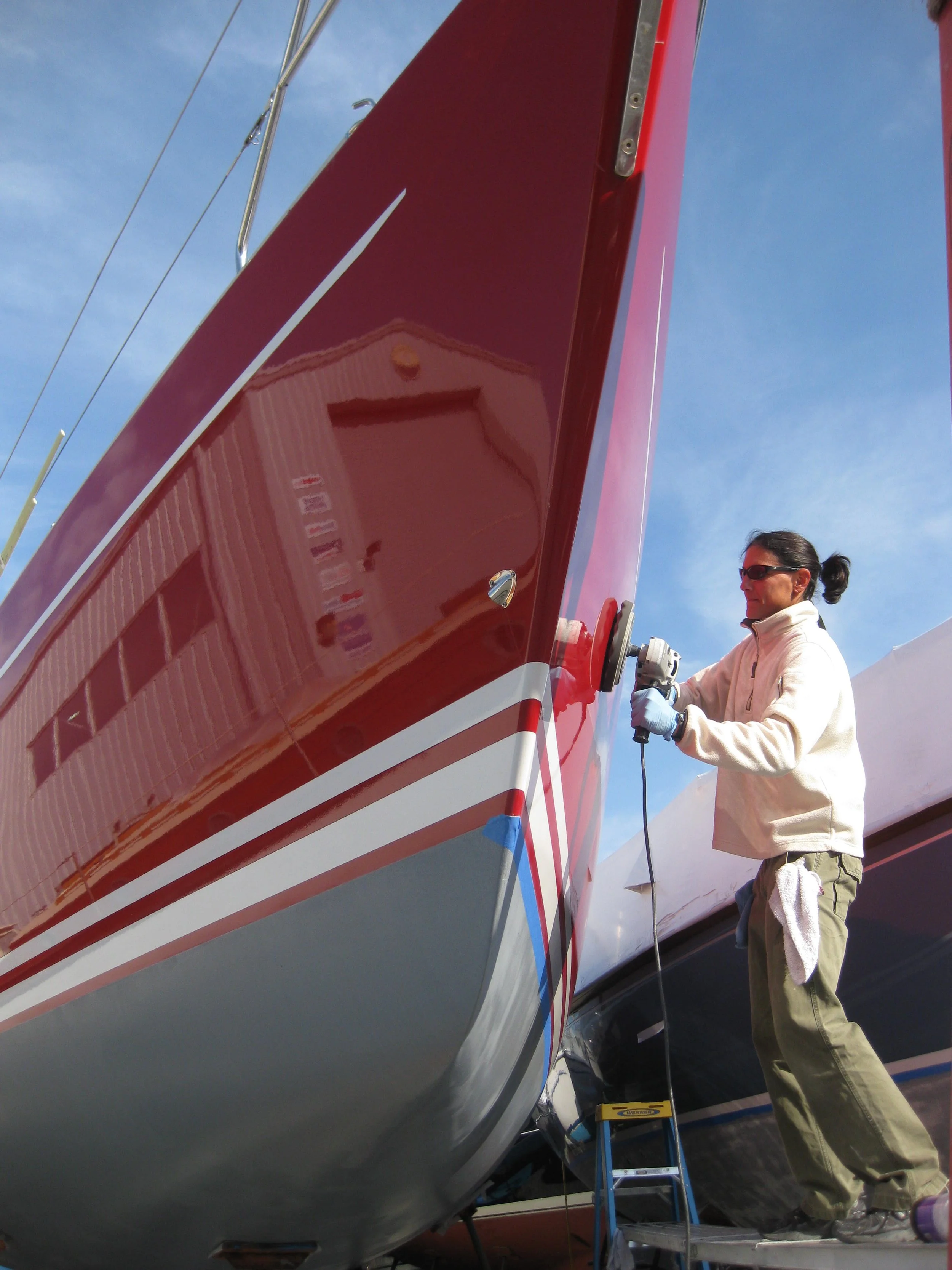 A person in a beige jacket and sunglasses is working on the side of a red and white sailboat, using a power tool on the hull. The boat is elevated, and the person is standing on a small ladder with a towel hanging from their pocket. The sky is blue with some clouds.