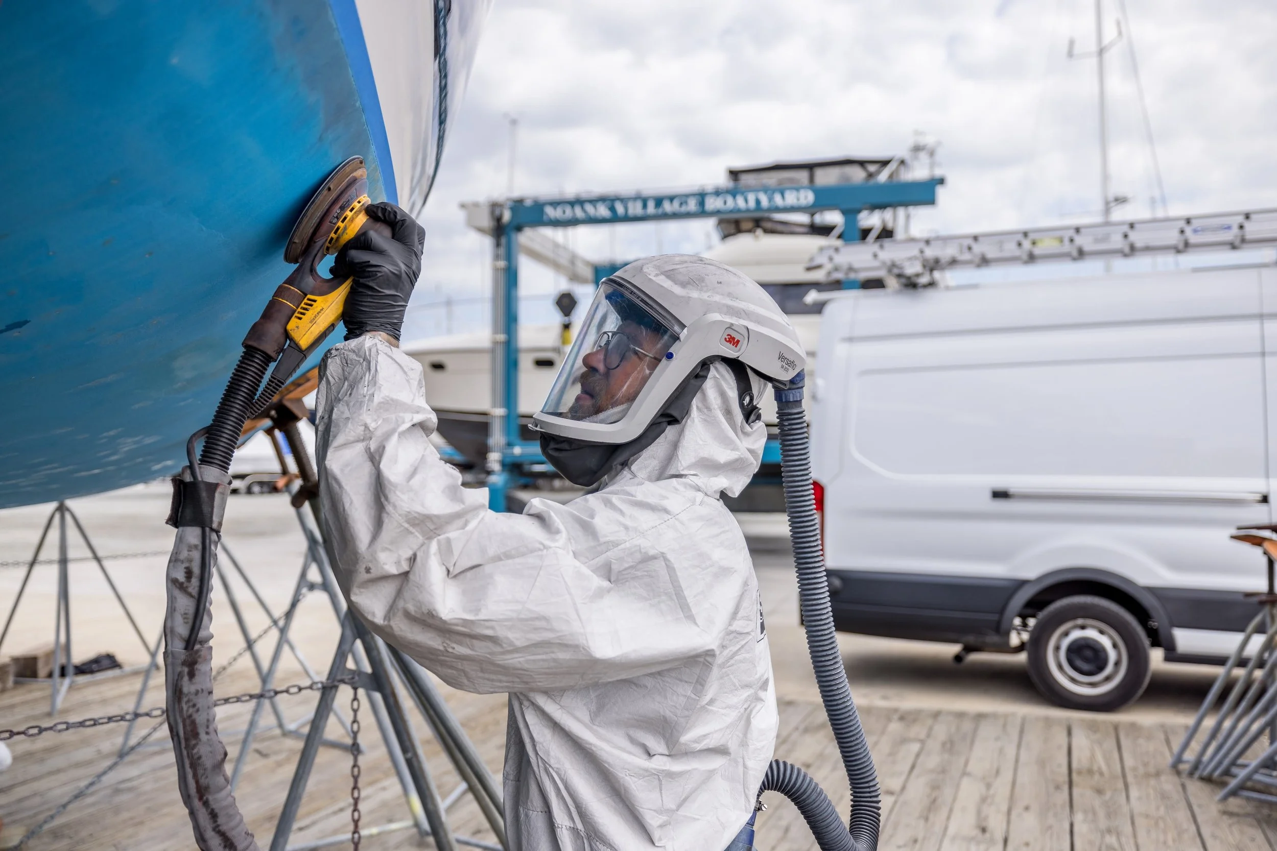 A person in protective gear, helmet, and gloves polishing the hull of a boat with a power buffer at a boat yard.