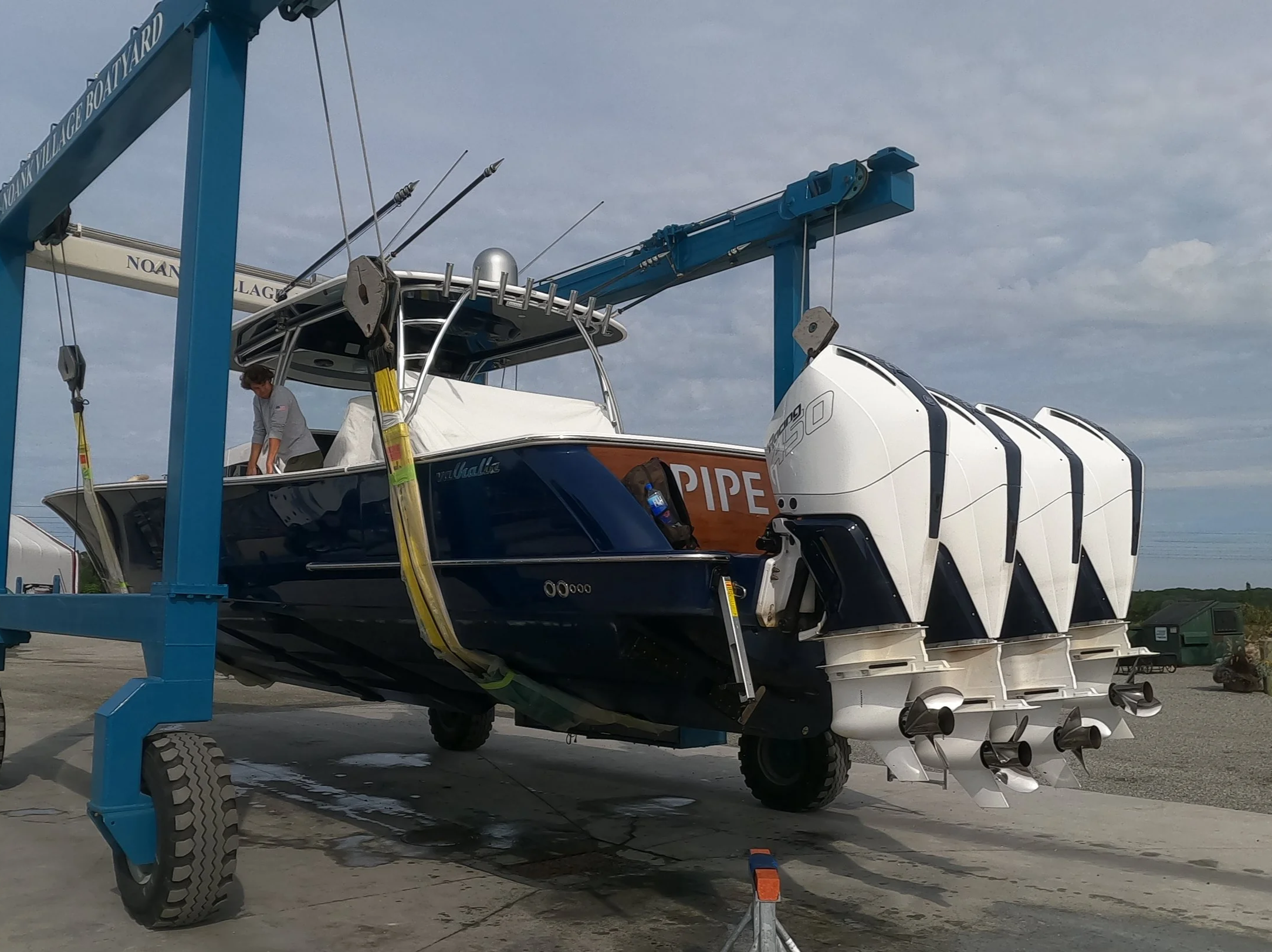 A boat is being lifted out of the water by a blue boat lift with a person onboard, standing near the boat's bow. The boat has a white and navy color scheme, and the outboard motor is visible at the stern. There is a cloudy sky above, and other boats and equipment are visible in the background.