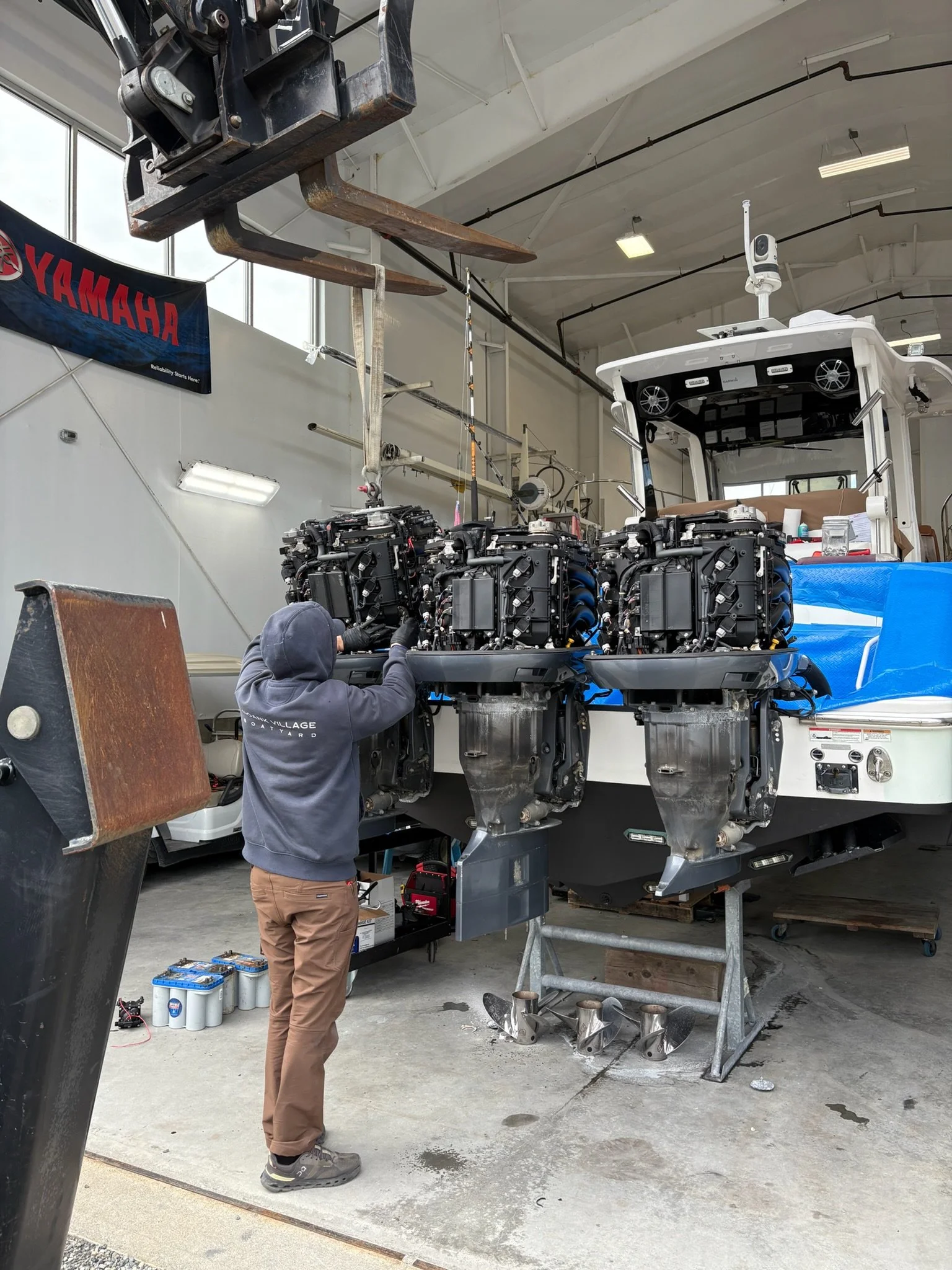 A person is working on an outboard motor inside a workshop. The motor is mounted on a stand, and the person is handling tools near it. The workshop has a high ceiling with fluorescent lights, a Yamaha banner, and various equipment and tools around.