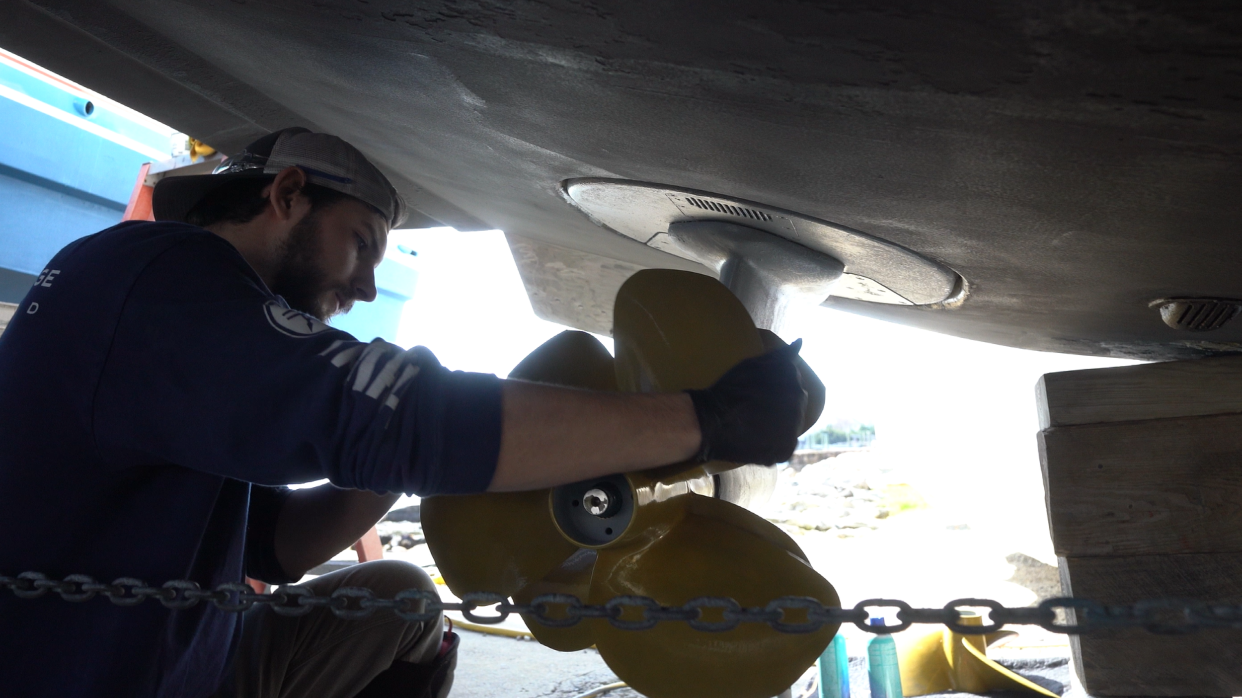 A man working underneath a boat, using a power tool to sand or smooth the hull of the boat.