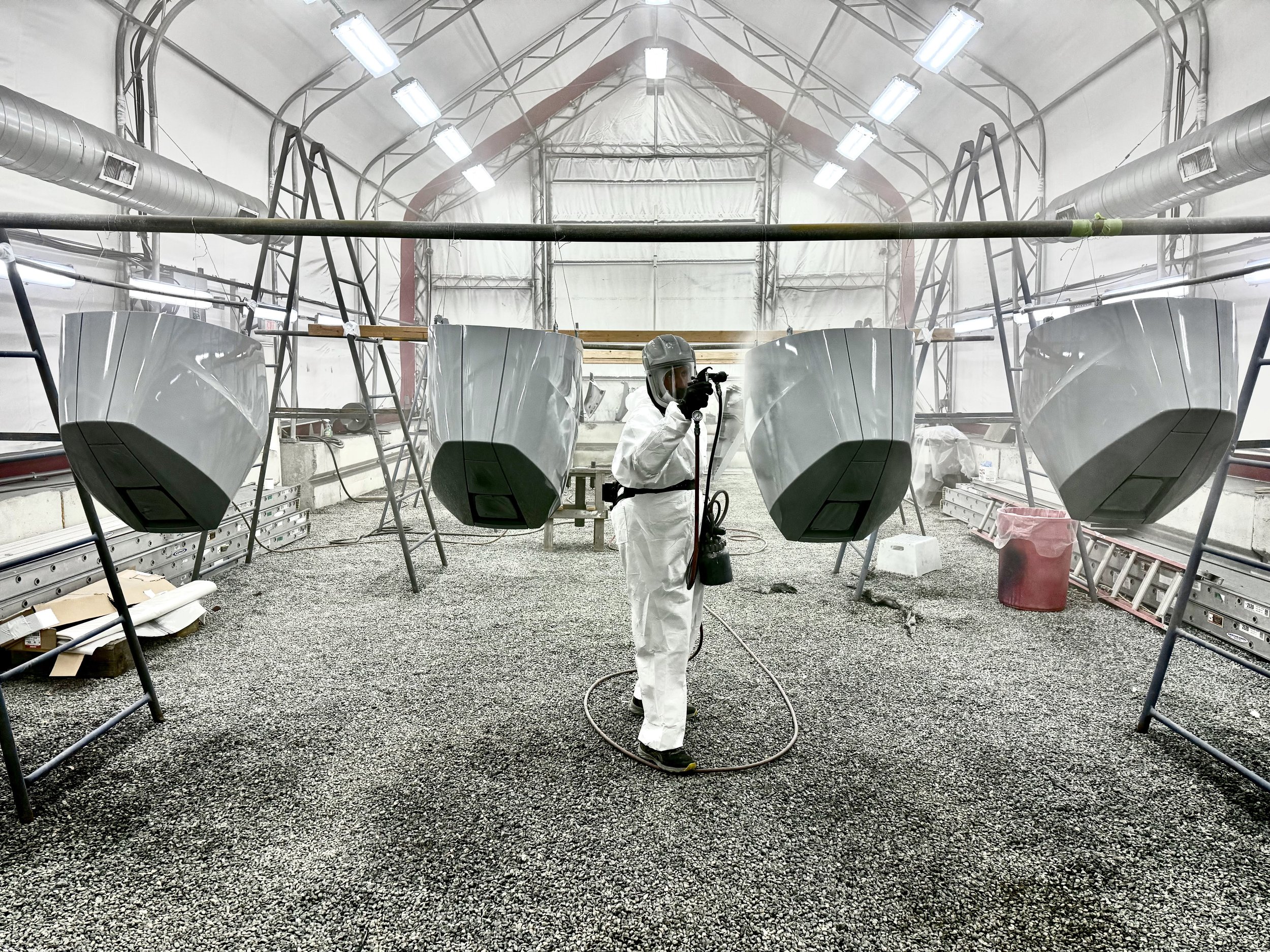 A person in protective gear, including a white suit, helmet, and mask, is spray painting large, white, streamlined objects inside a spacious, well-lit industrial tent with scaffolding and ventilation pipes.