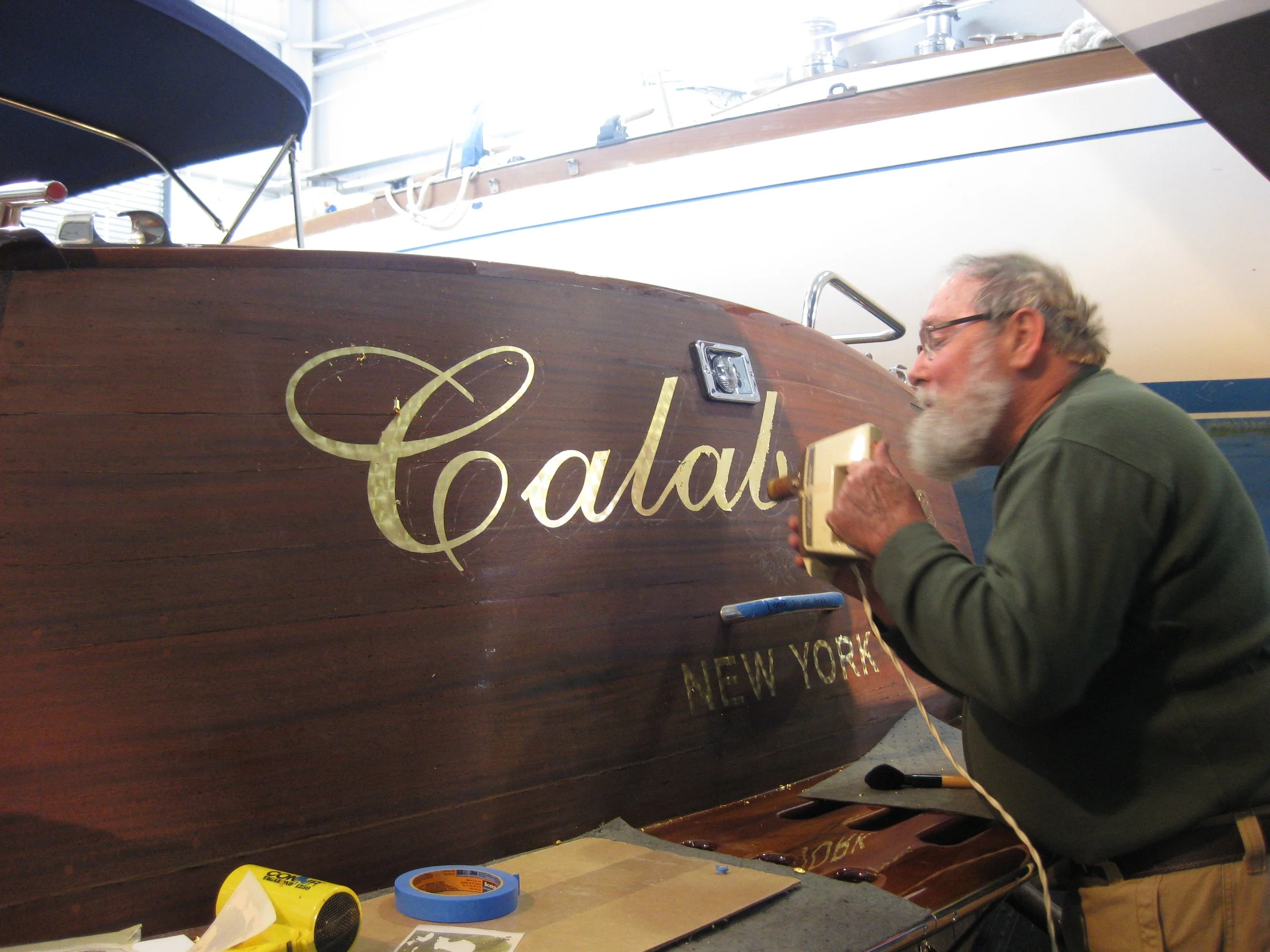 A man with glasses and a beard is using gold lettering to decorate a wooden boat, with tools and tape visible on a workbench under bright natural light.
