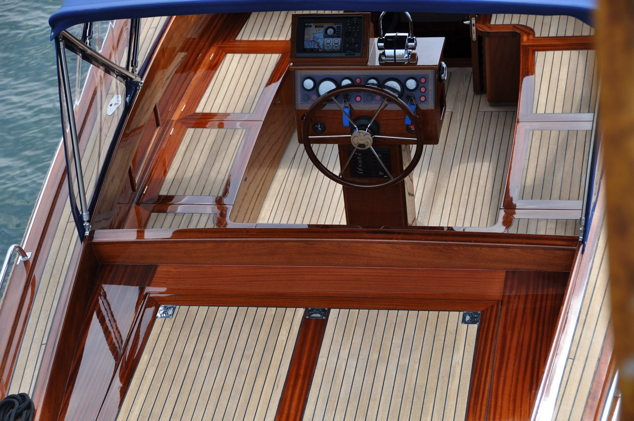 The helm of a wooden boat with navigation equipment and a steering wheel on a striped wood deck.