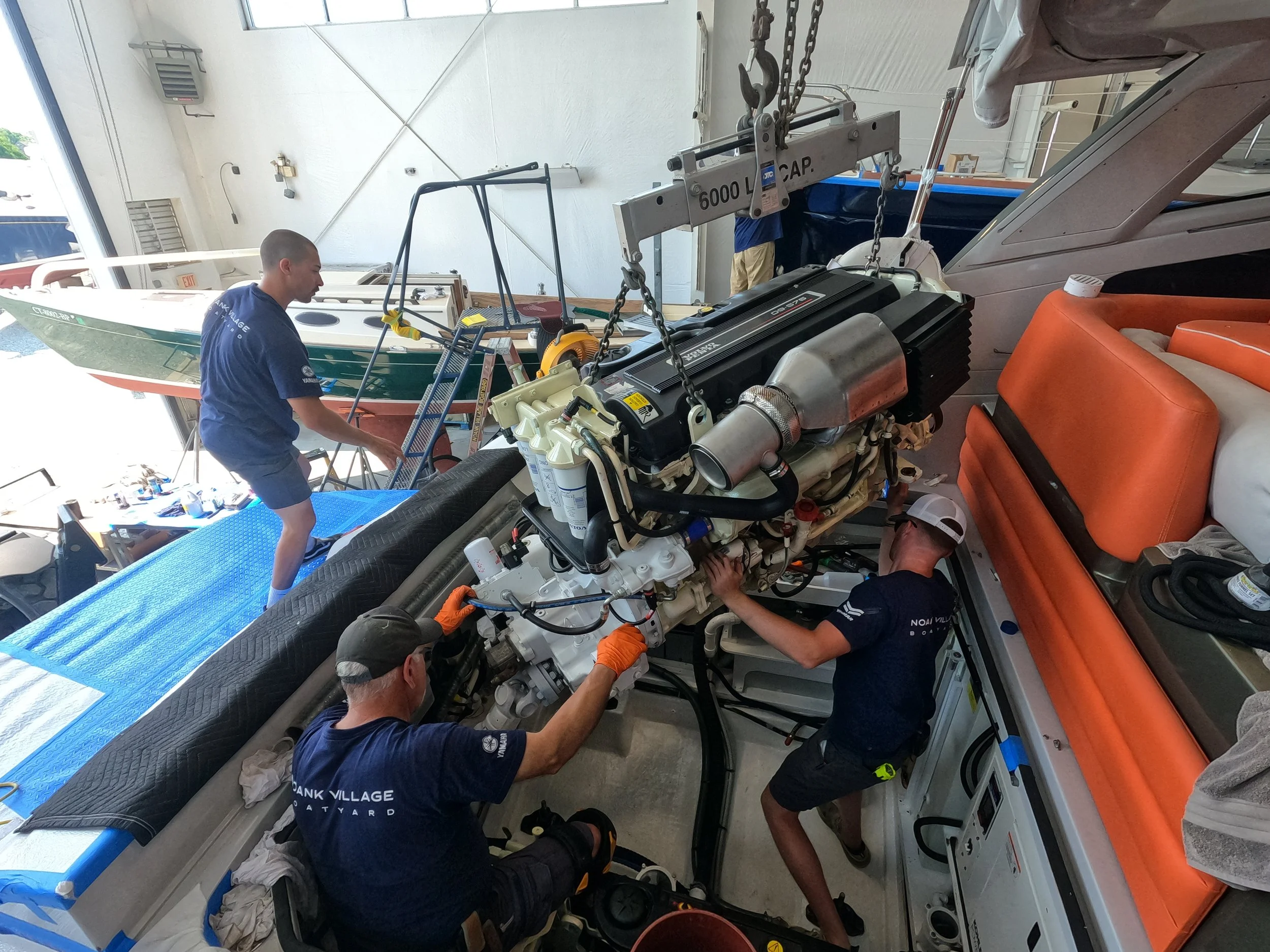 A group of workers inside a boat storage area are installing or repairing a boat engine. They are using tools while standing and kneeling around the engine, which is supported by a lift labeled '6000 LBS CAP.' The boat is on a trailer, and other boats and equipment are visible in the background.