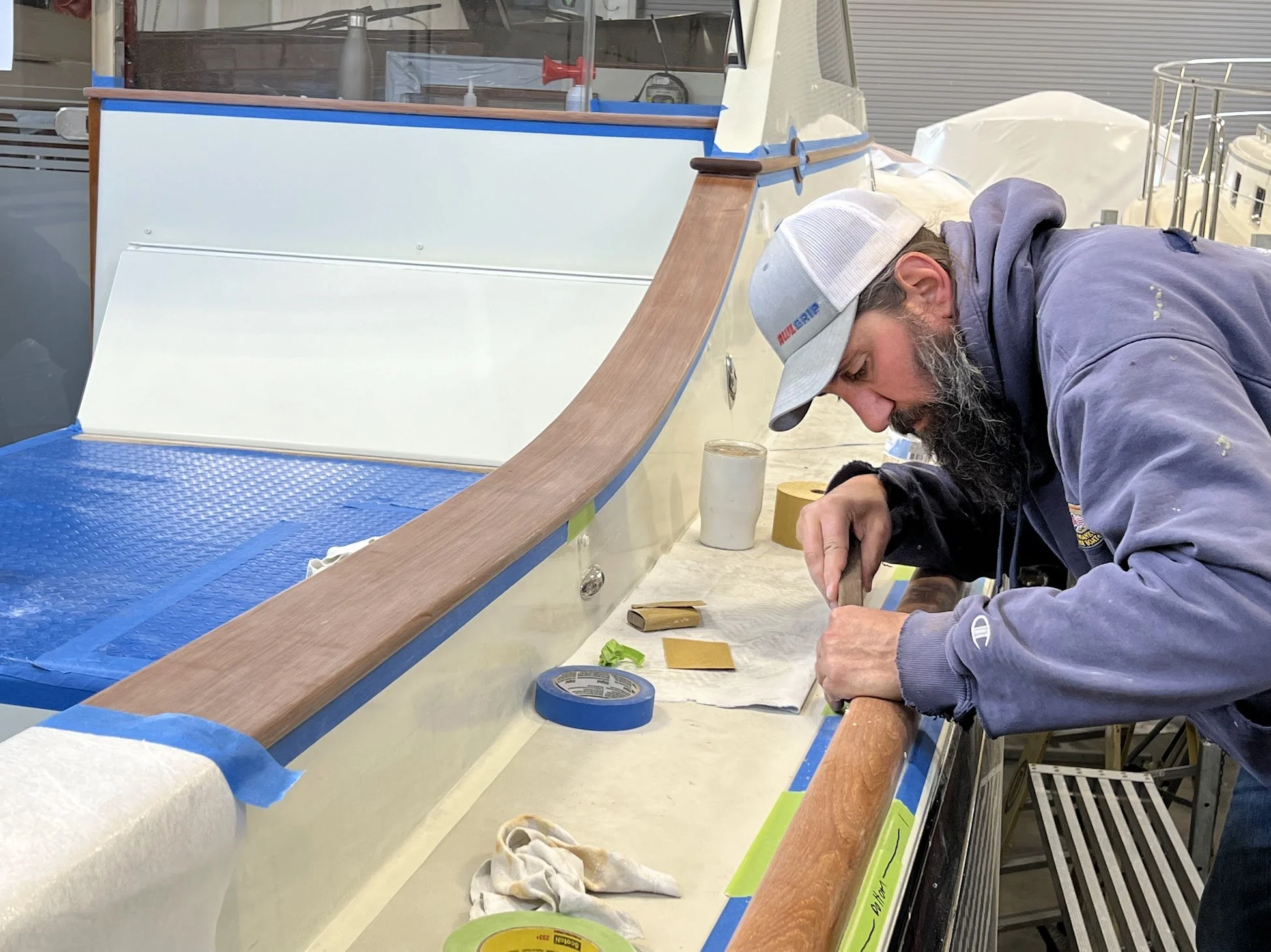 A man with a beard wearing a grey cap and purple hoodie working on a boat in a workshop. The boat has a wooden trim and a white interior, with tools and tape around him.