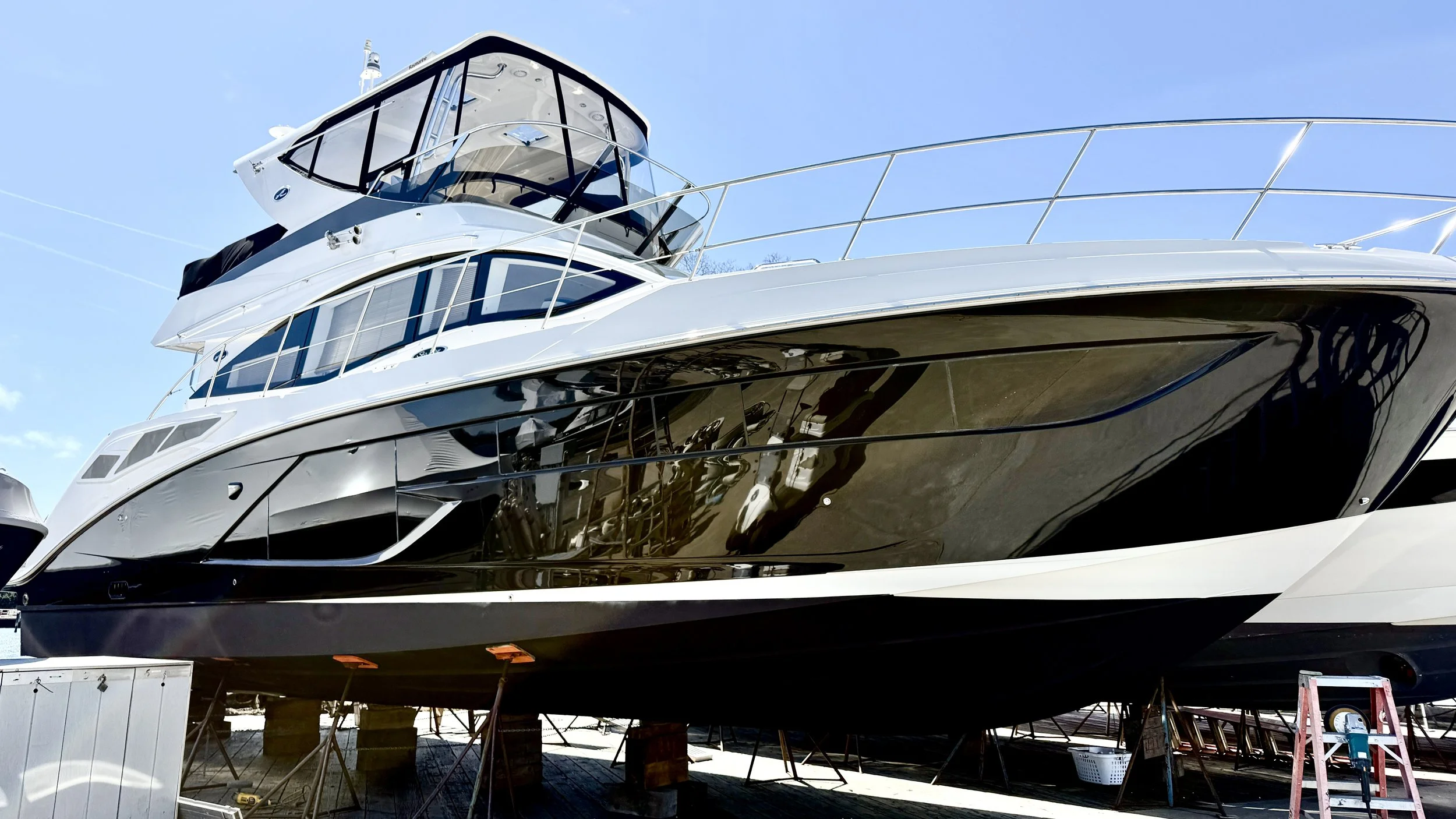 Large modern yacht on land with supports underneath, reflection of the boat in the black hull, blue sky in the background.