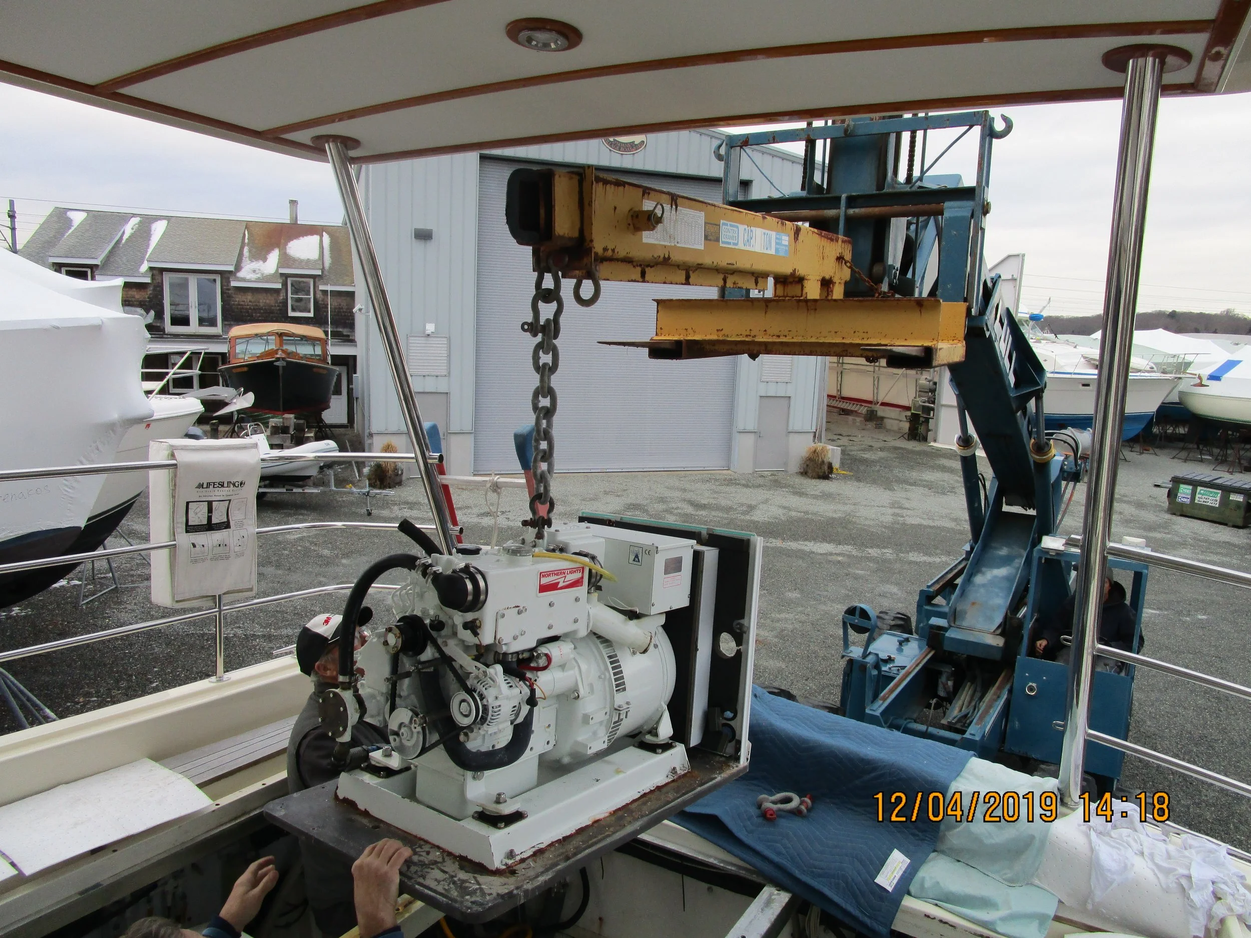 A large white machine with motor components on a table, connected to a pipeline, at an outdoor boatyard. Multiple boats are stored on land in the background, with a metal building and cloudy sky overhead.