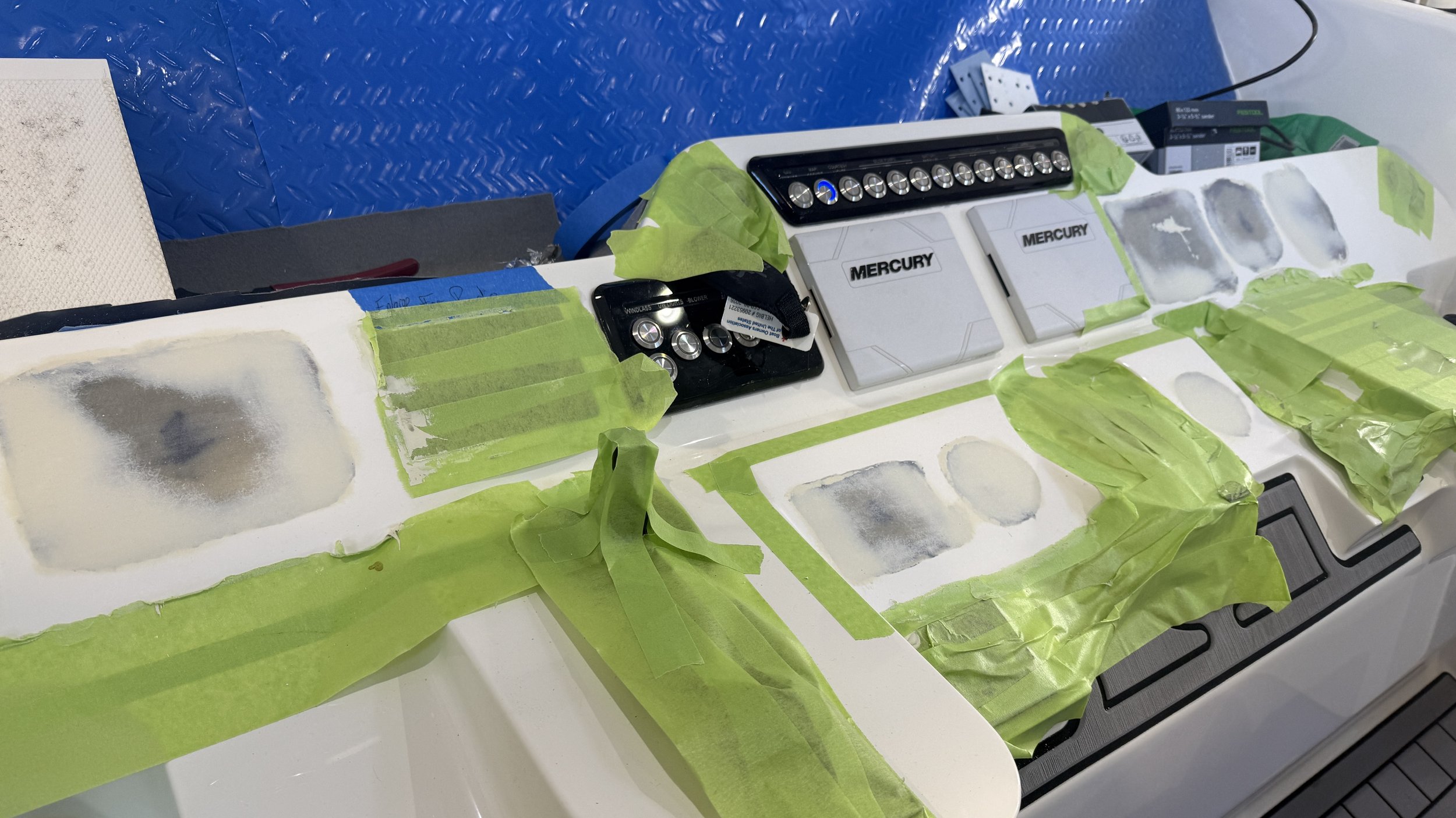 A workbench with multiple sections covered in painter's tape and primer, with various tools and electronic components including a black device with buttons and switches, and boxes labeled Mercury.