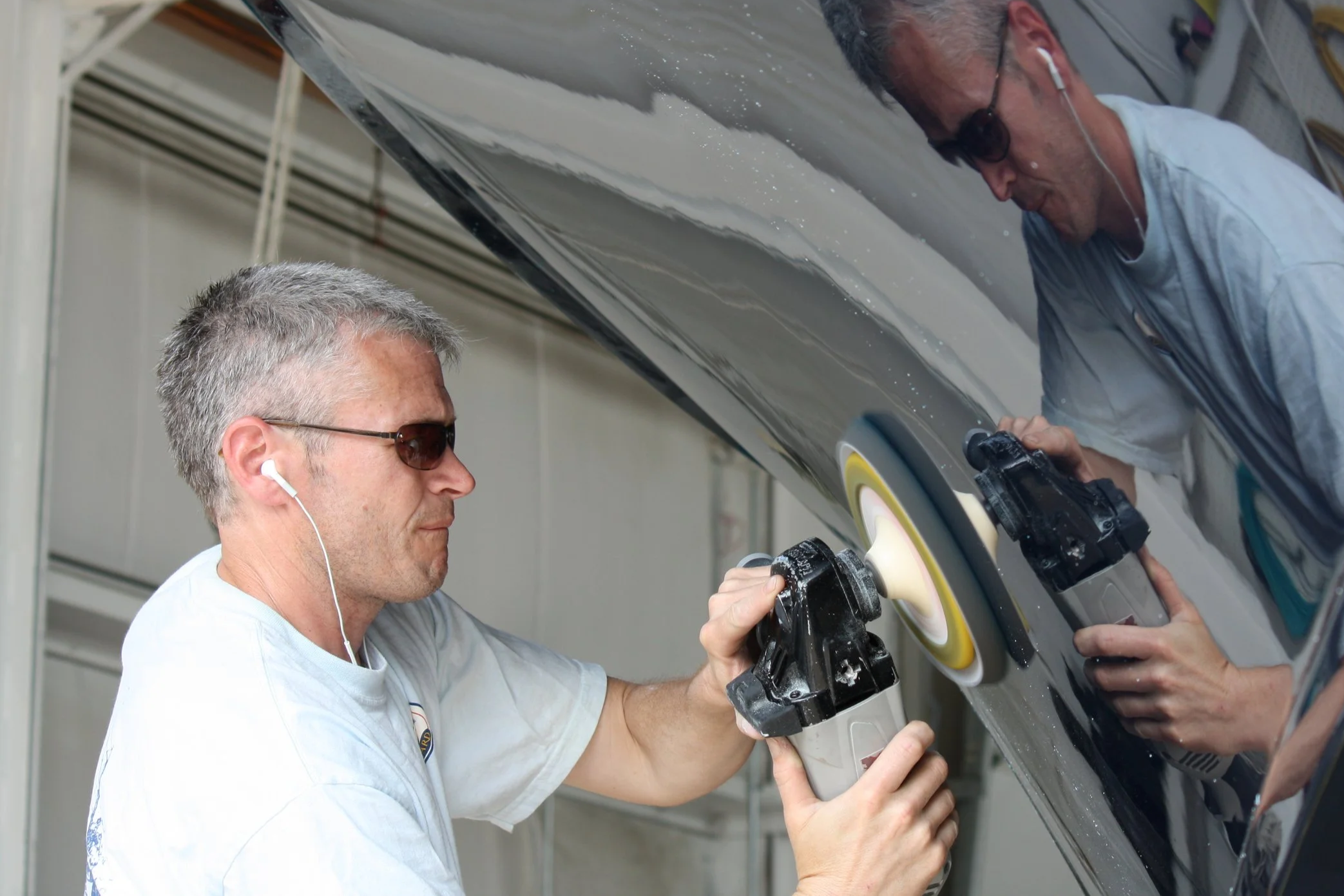 Two men working on polishing a car's body, with one man using a power buffer and the other holding a tool, both wearing white shirts, sunglasses, and earphones.