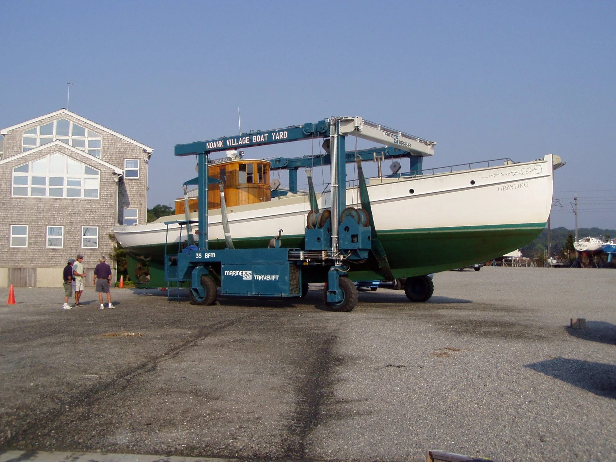 A boat named 'Grayling' is being hoisted by a blue crane at a boat yard, with a group of people observing nearby, in front of residential buildings under a clear sky.