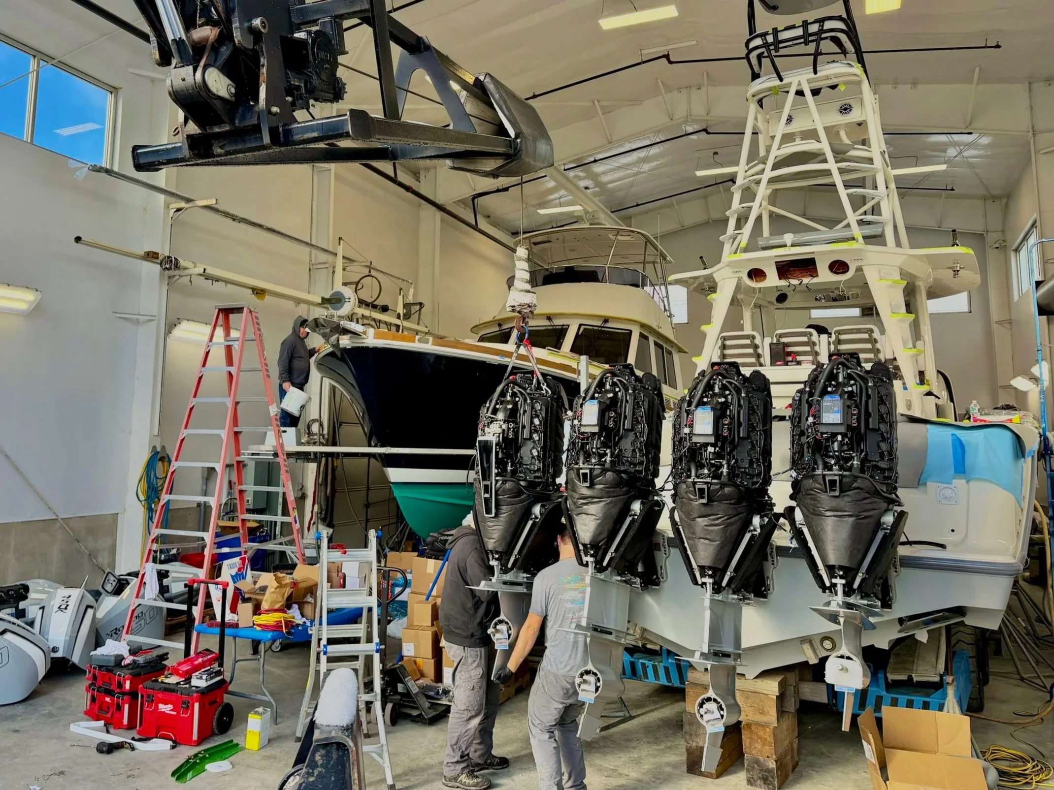 Workers assembling a boat with multiple outboard engines in a factory. The boat is suspended, and various tools, boxes, and equipment are scattered around.