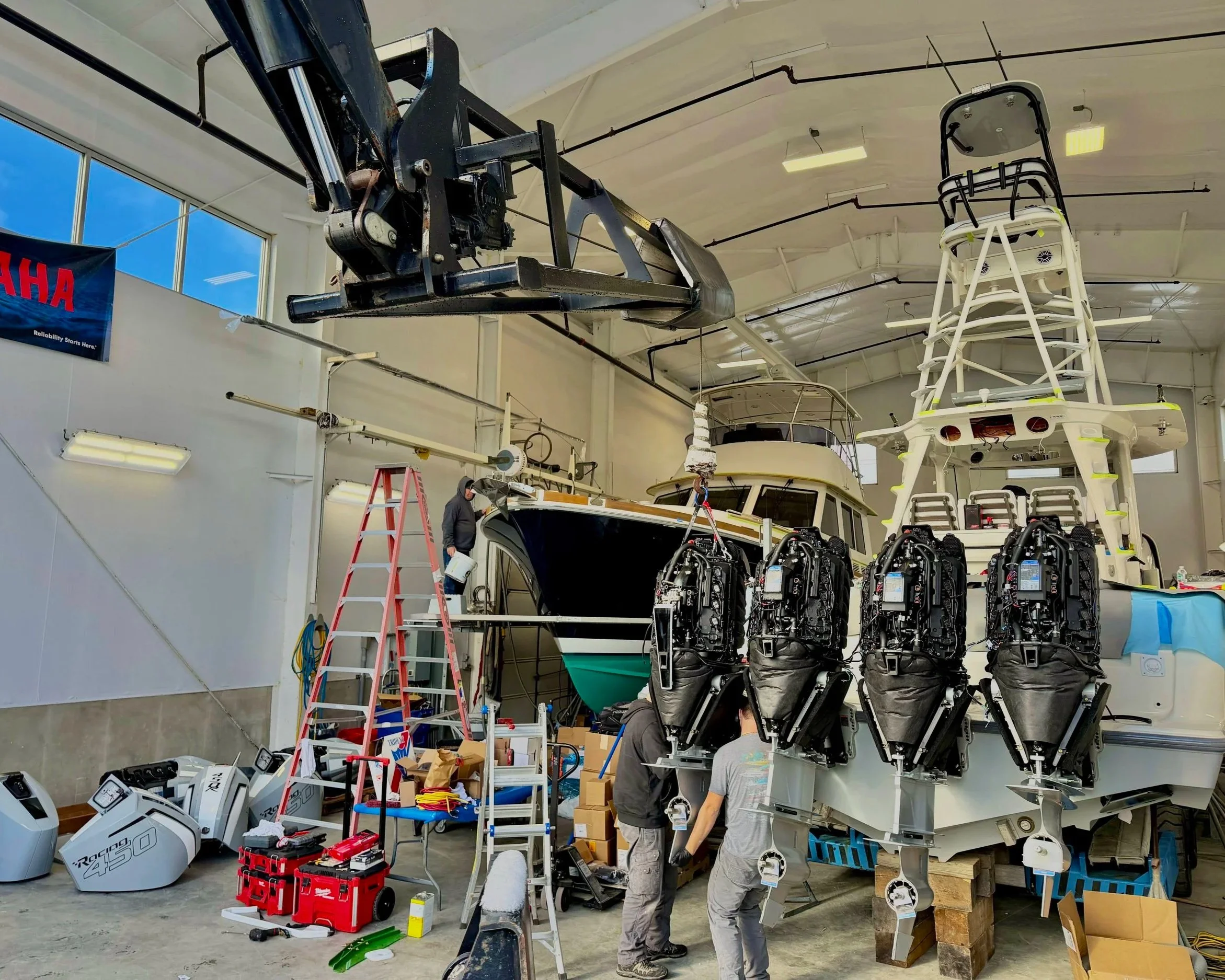 Inside a workshop with boat parts and equipment hanging and stacked, workers are assembling or repairing boat engines and structures, surrounded by tools, ladders, and boxes.