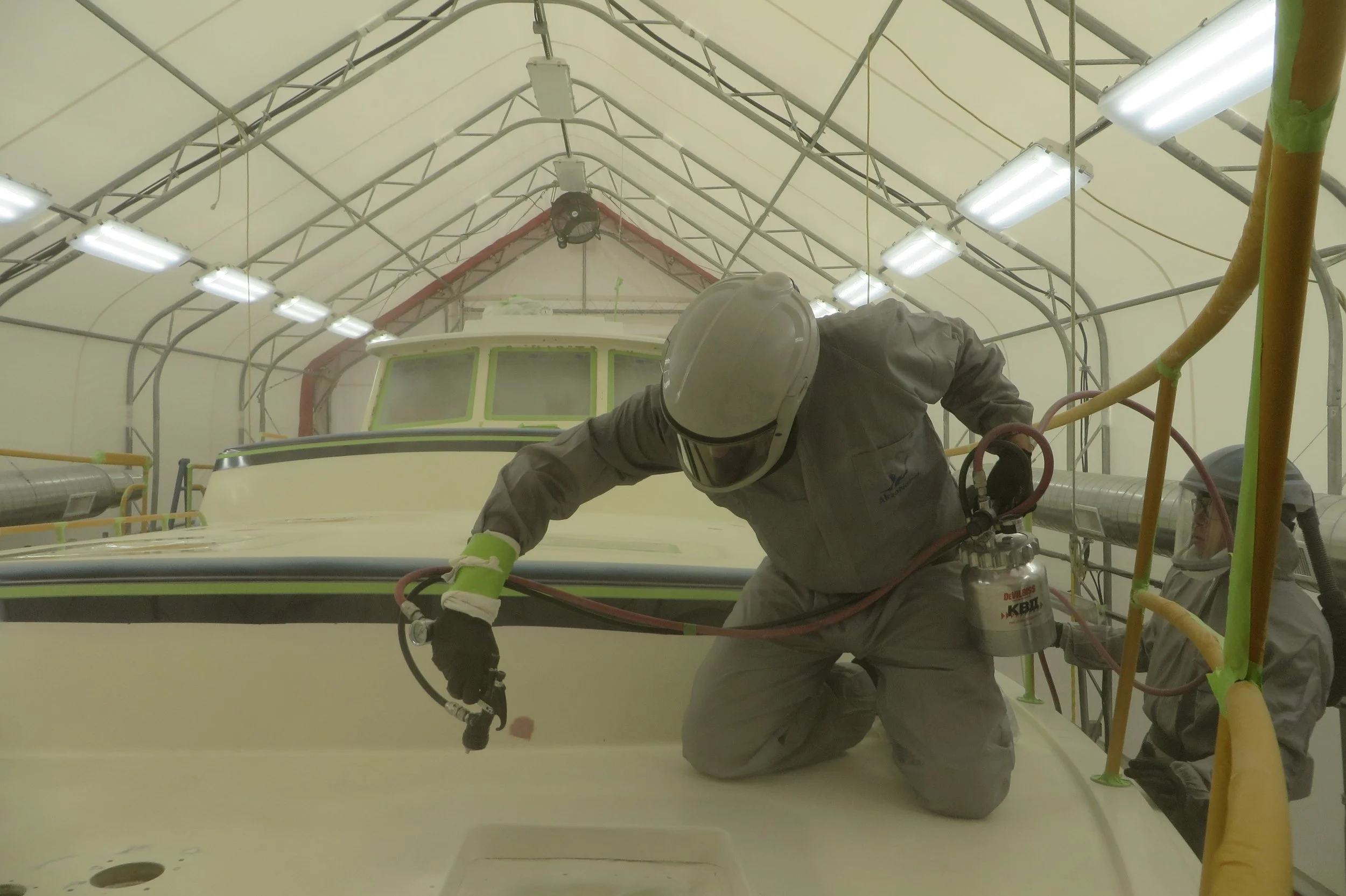 A worker wearing protective gear, including a helmet and gloves, is kneeling on the floor inside a large, brightly lit, industrial enclosure, using equipment to work on the surface of a large vehicle or structure.