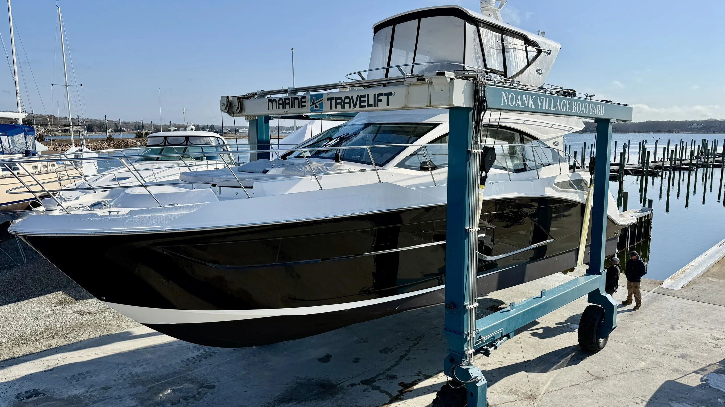 A large yacht is on a boat lift at a marina, with other boats docked nearby and calm water in the background.
