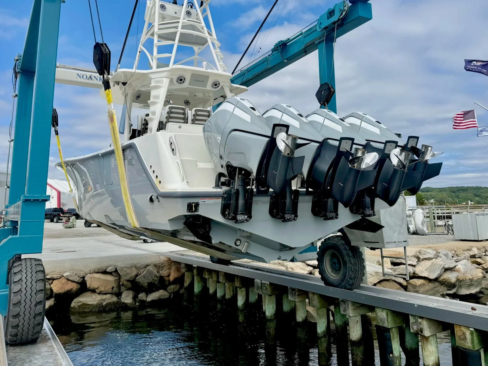 A large white boat with multiple outboard motors parked on a trailer under a blue crane, near a waterway with rocks and a wooden dock.