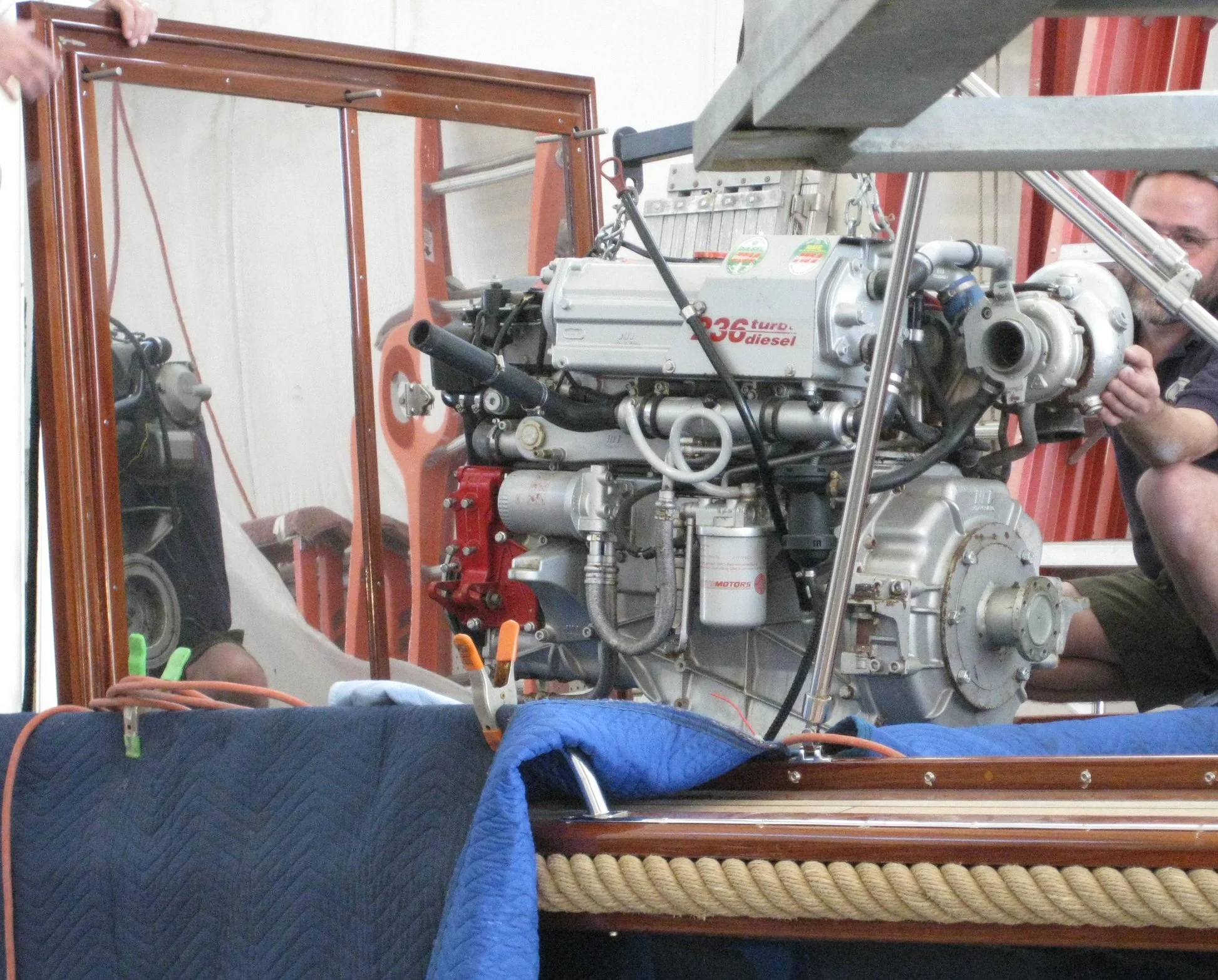 View of a man working on a turbocharged diesel engine inside a wooden framed structure, with tools, a mirror, and a blue blanket or cover in the foreground.