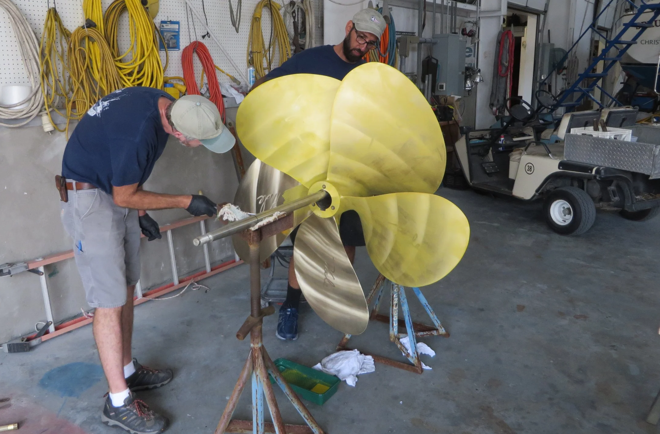 Two men working on a large yellow metal flower sculpture in a workshop, surrounded by tools and equipment.