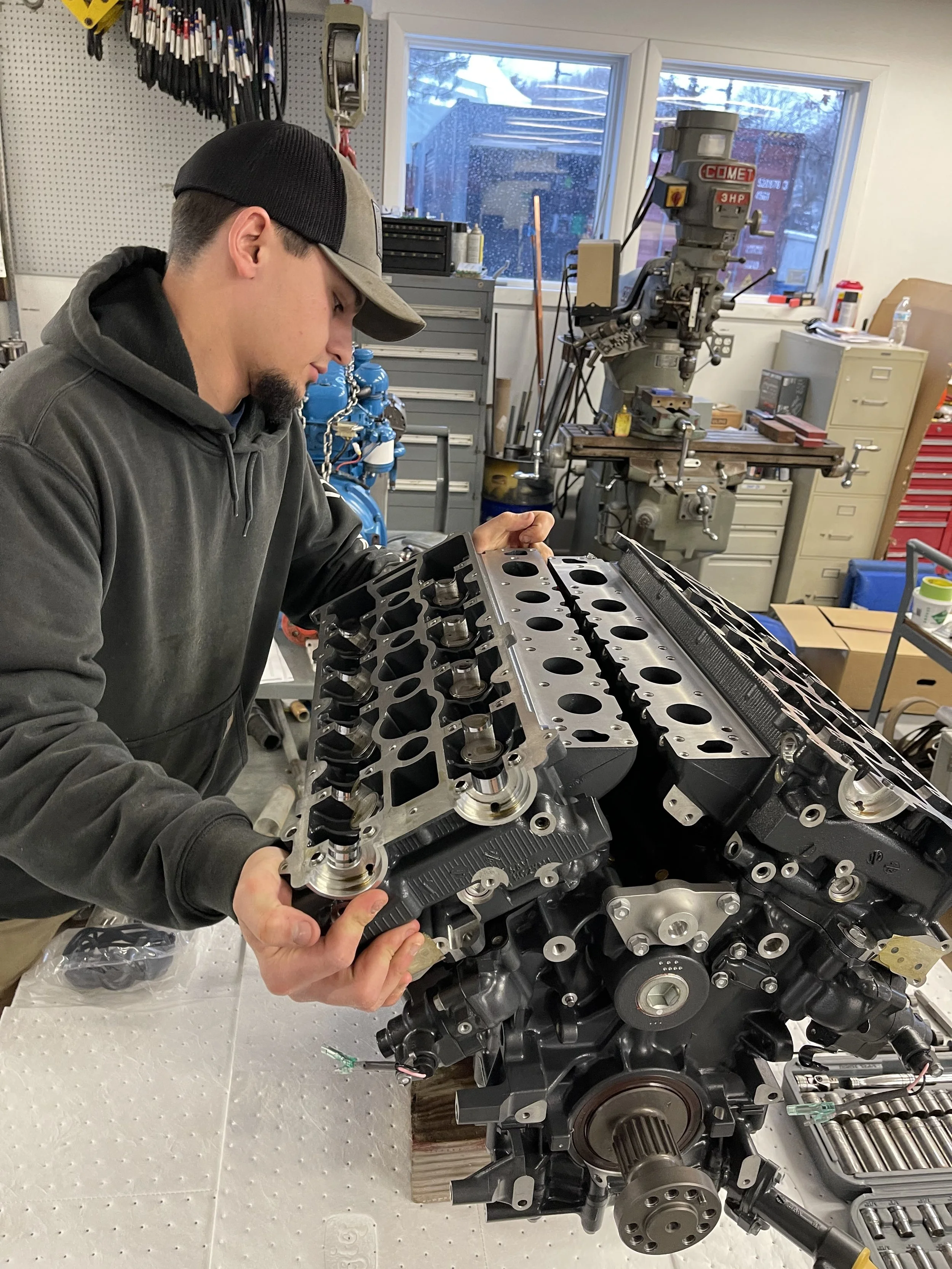 A person in a workshop holding an engine cylinder head over a partially assembled engine block with various tools and equipment around.