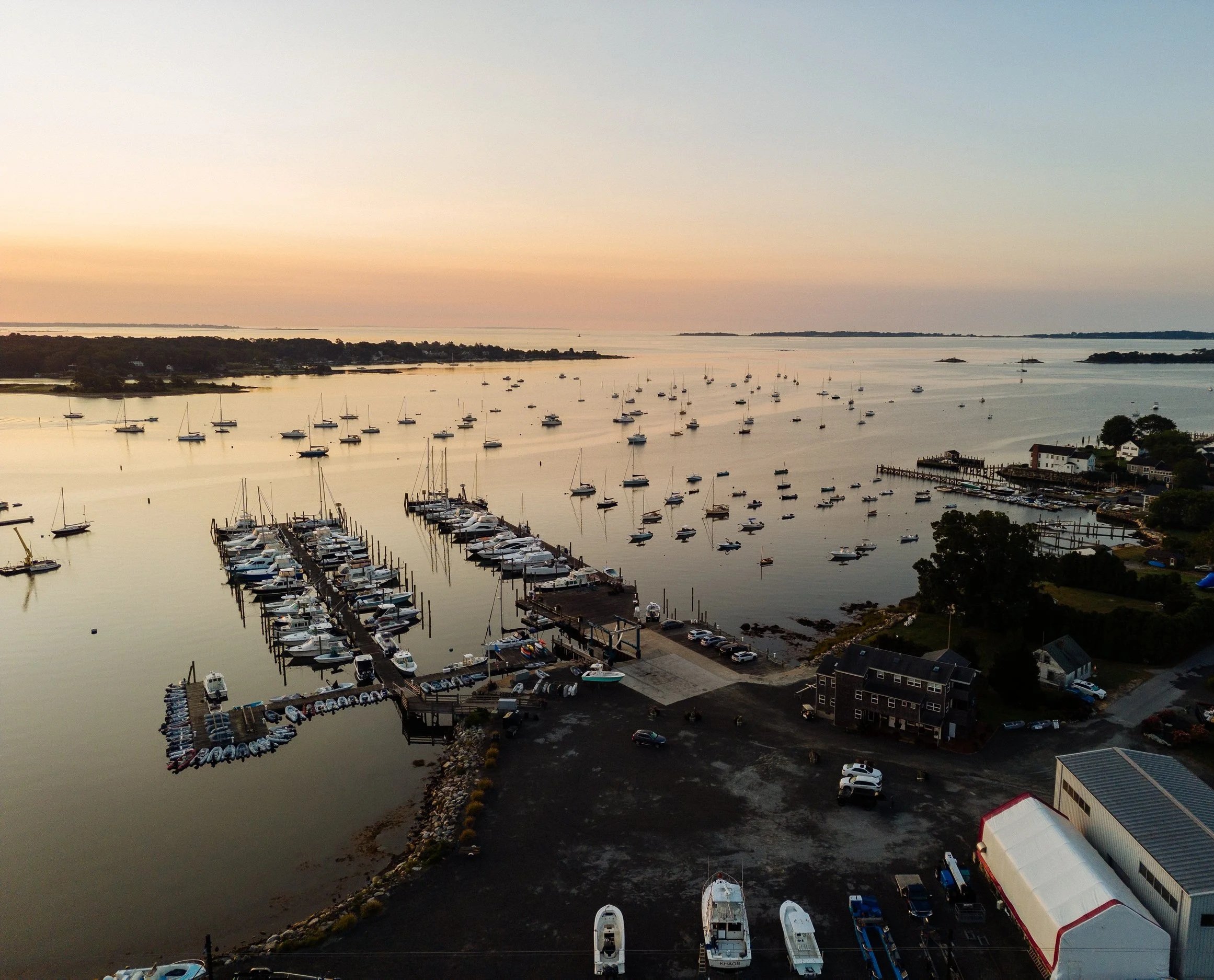 Aerial view of a harbor with numerous boats and yachts docked along piers at sunset, with houses and trees lining the shore, and a calm, reflective water surface.
