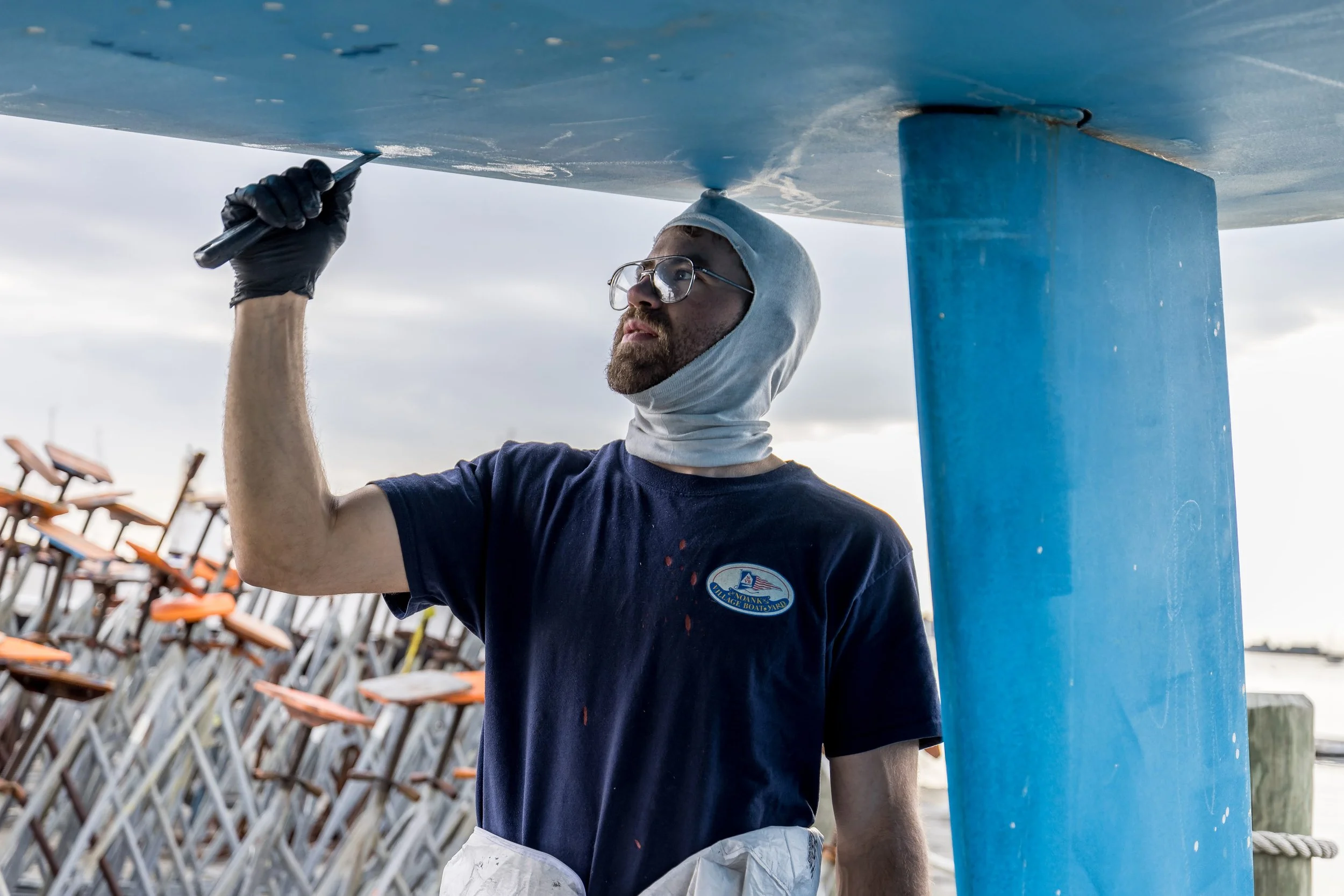 A man with glasses and a hoodie repairing or inspecting a blue boat underwater during the daytime. Snowy landscape in the background.