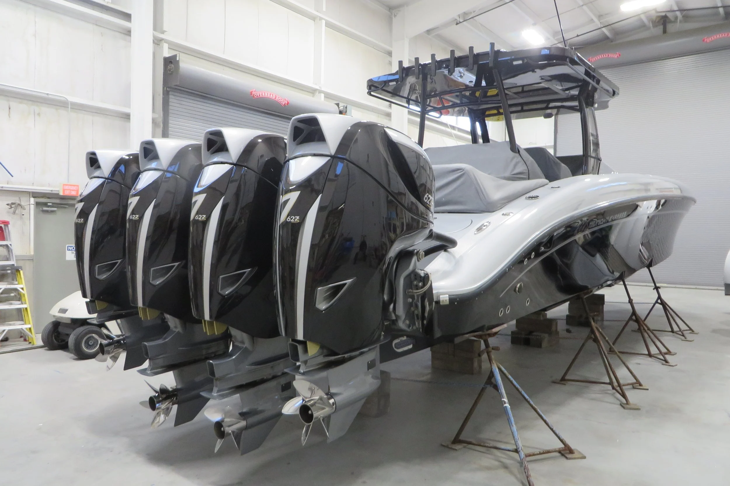 A large modern boat with five outboard motors attached at the back, stored indoors on a platform supported by wooden blocks.