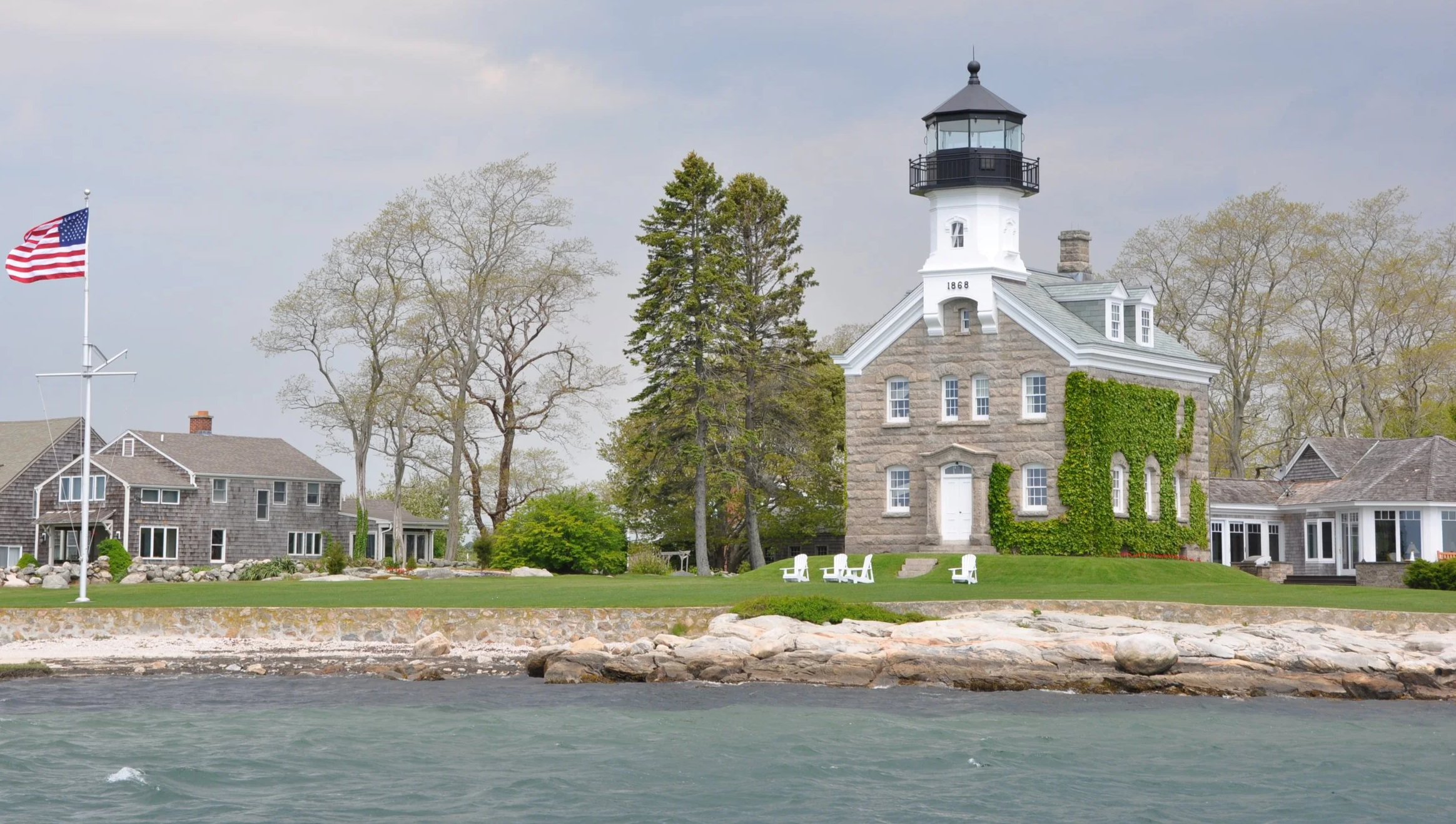 Lighthouse with a black top and white base, built in 1868, surrounded by trees and houses, on a coastline with rocks and water in the foreground.