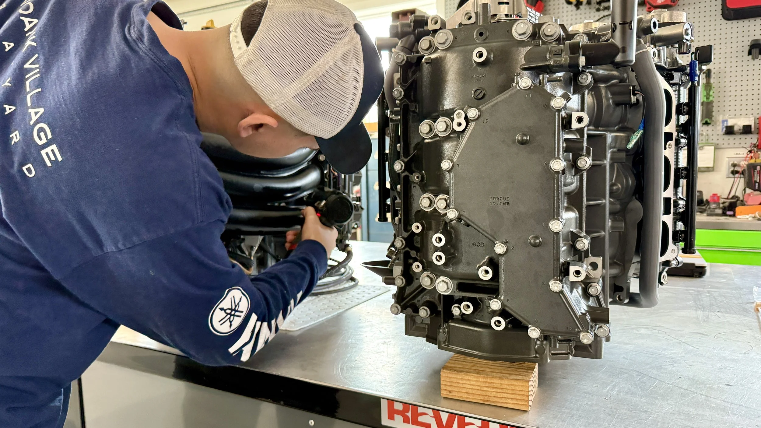 A person wearing a blue shirt, gray mesh cap, and glasses working on a large mechanical engine or machine in a workshop. The person is using a tool on the equipment, which is secured to the table with wooden blocks.
