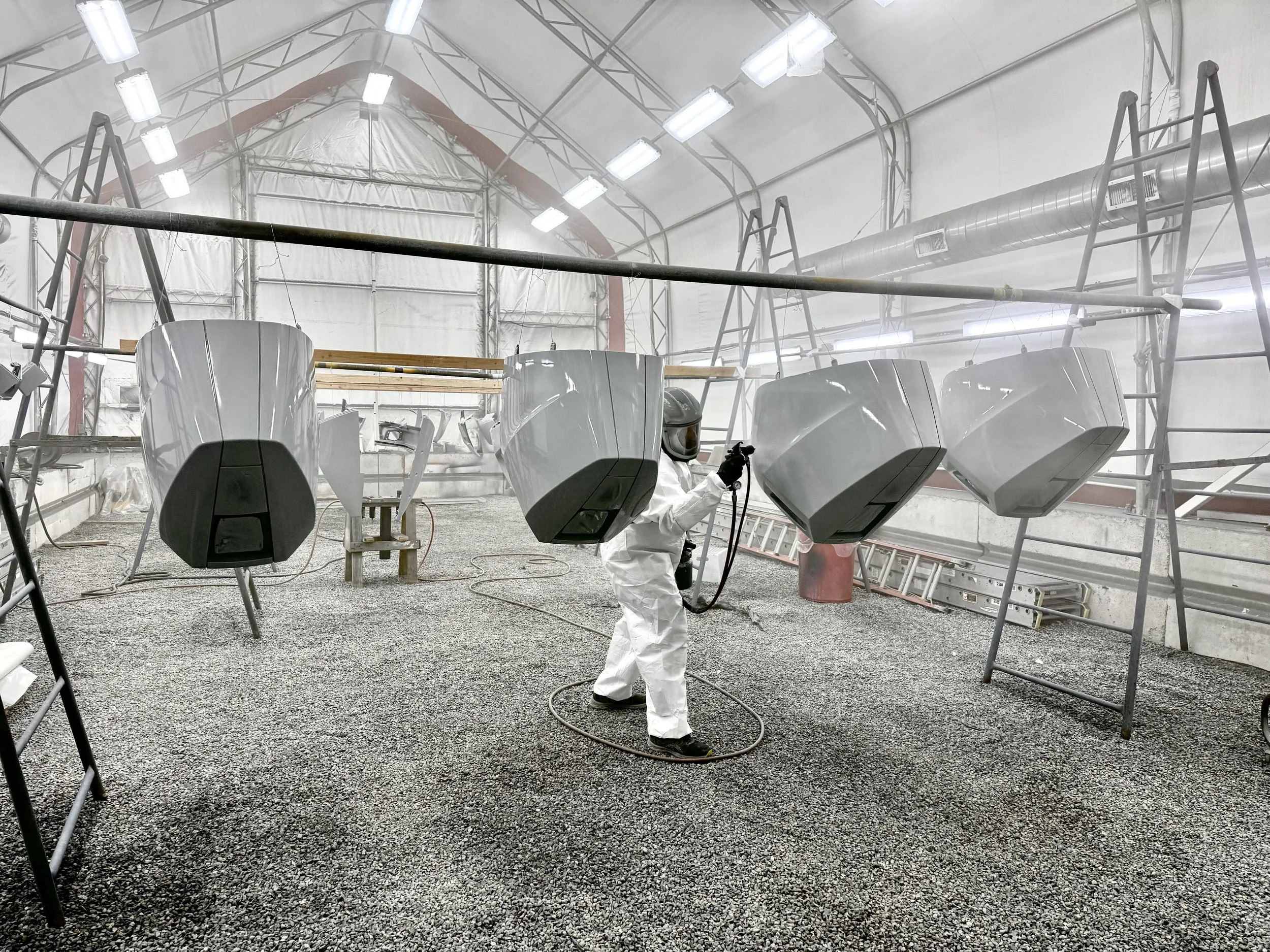 A person in white protective coveralls and a helmet, holding a spray gun, standing inside an industrial or manufacturing workshop. Several large objects are hanging from the ceiling, and there is scaffolding and ladders around the area. Bright overhead lights illuminate the space.
