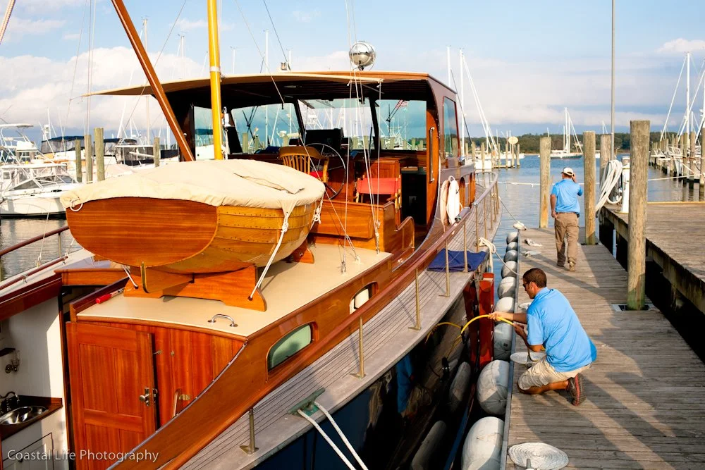 Two men are cleaning or inspecting a wooden yacht docked at a marina, with one kneeling and the other walking on the pier, surrounded by sailboats and marina posts.