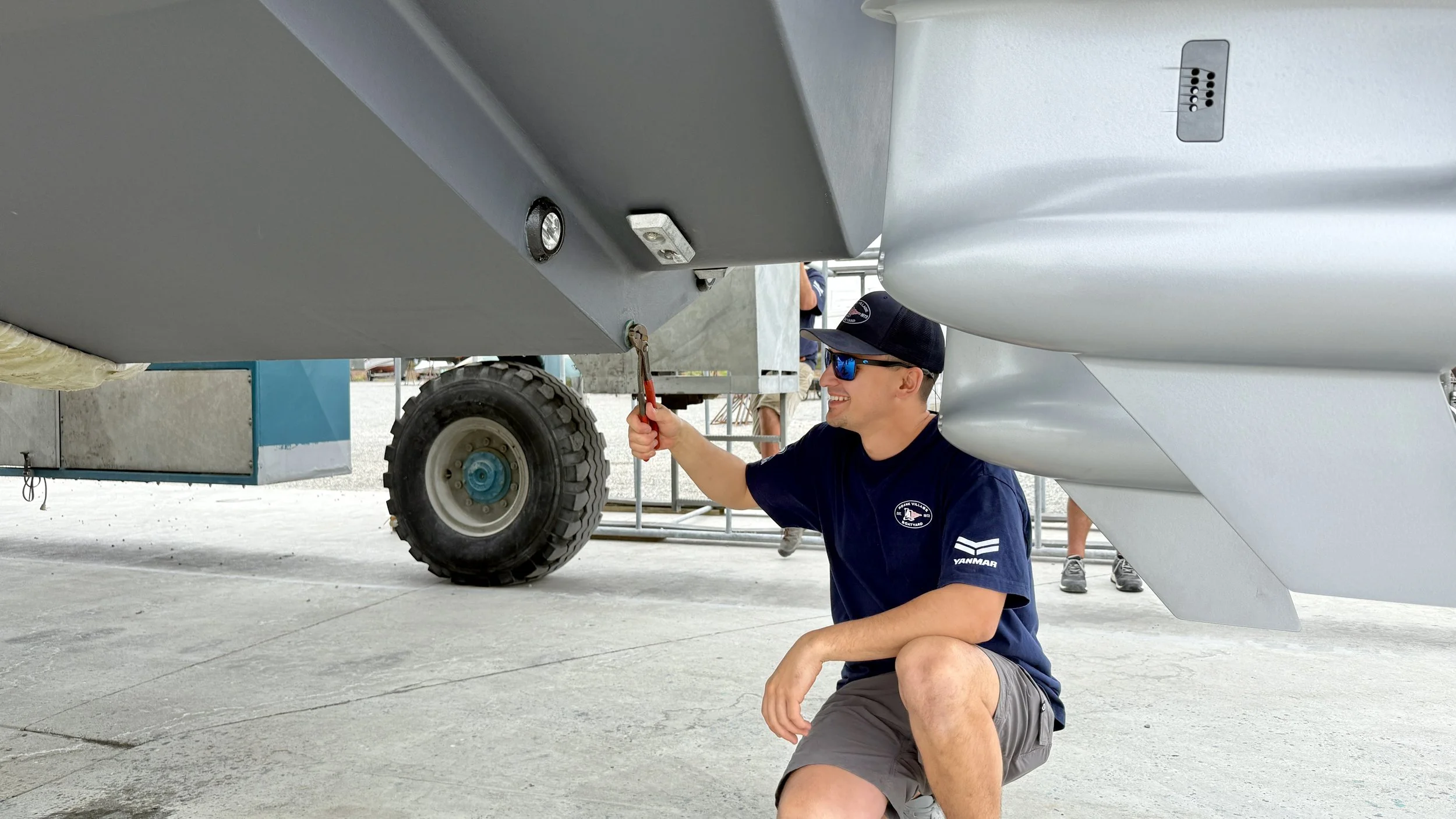 A man laughing and kneeling under an aircraft fuselage, working on or inspecting the underside with a wrench, while another person is seen in the background. The man is wearing sunglasses, a baseball cap, and a navy blue T-shirt with a logo.