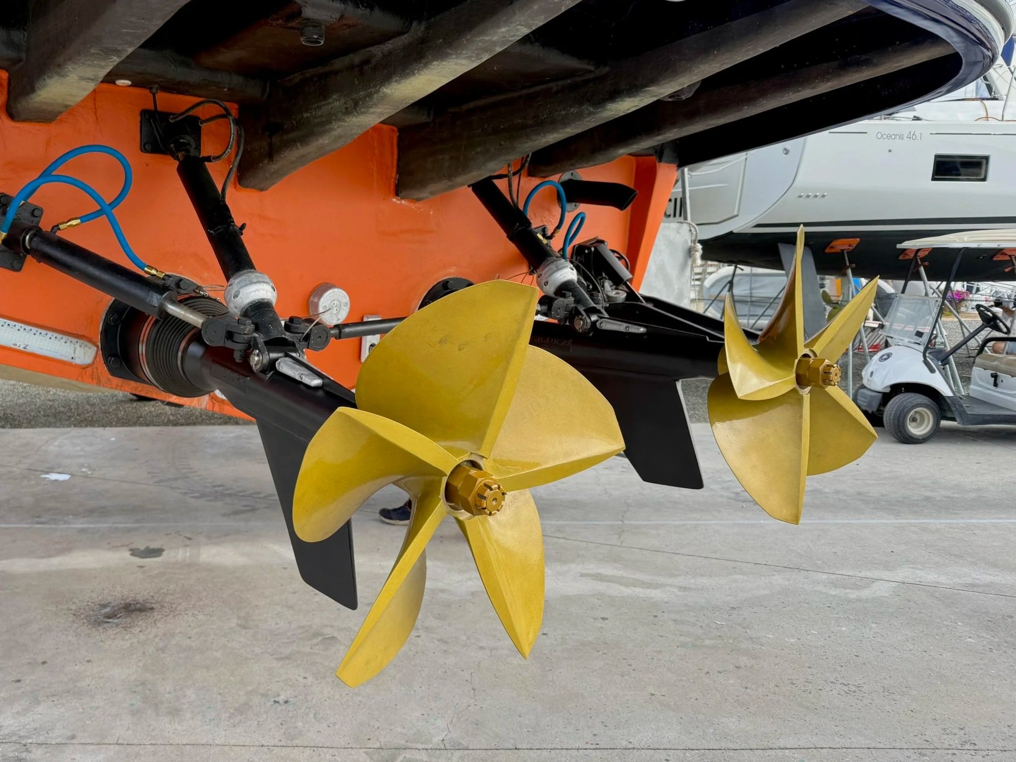 Close-up of a boat's propeller and mechanical components under the hull, with other boats and a golf cart in the background.