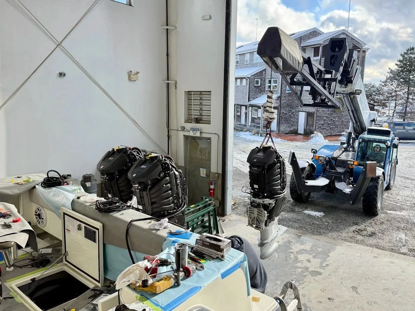 Inside a workshop with two robotic engines on workbenches and a lift holding large engine parts outside. In the background, buildings and snow-covered ground are visible.