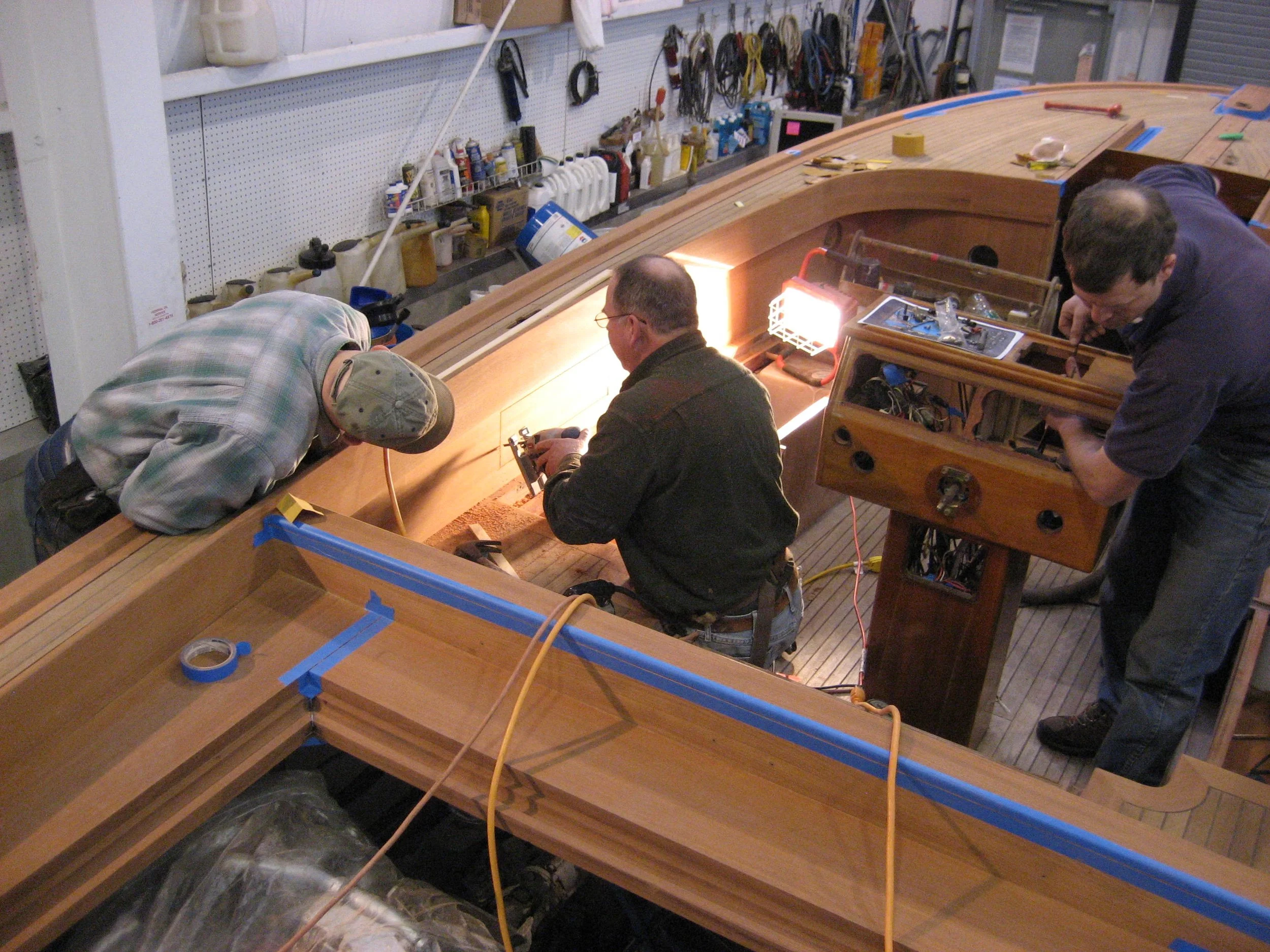 Three men working on a wooden boat in a workshop, surrounded by tools and supplies.