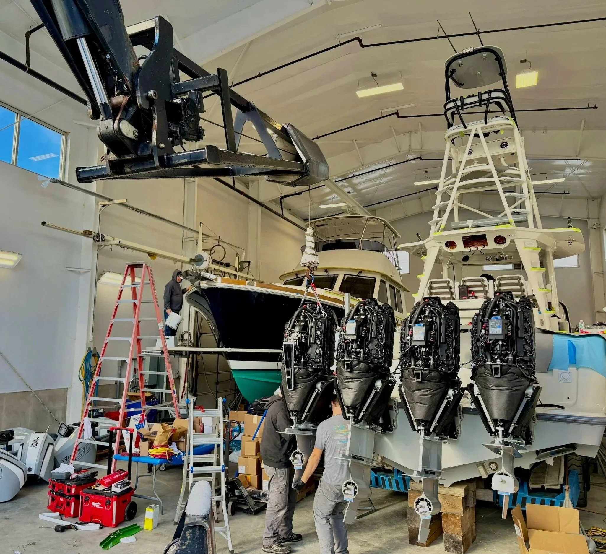 Several men are working on assembling or repairing boats or boat parts inside a large industrial workshop. There are multiple boats, tools, and equipment visible, including a large black boat hull on a stand, and some boat motor engines hanging in front of the men.