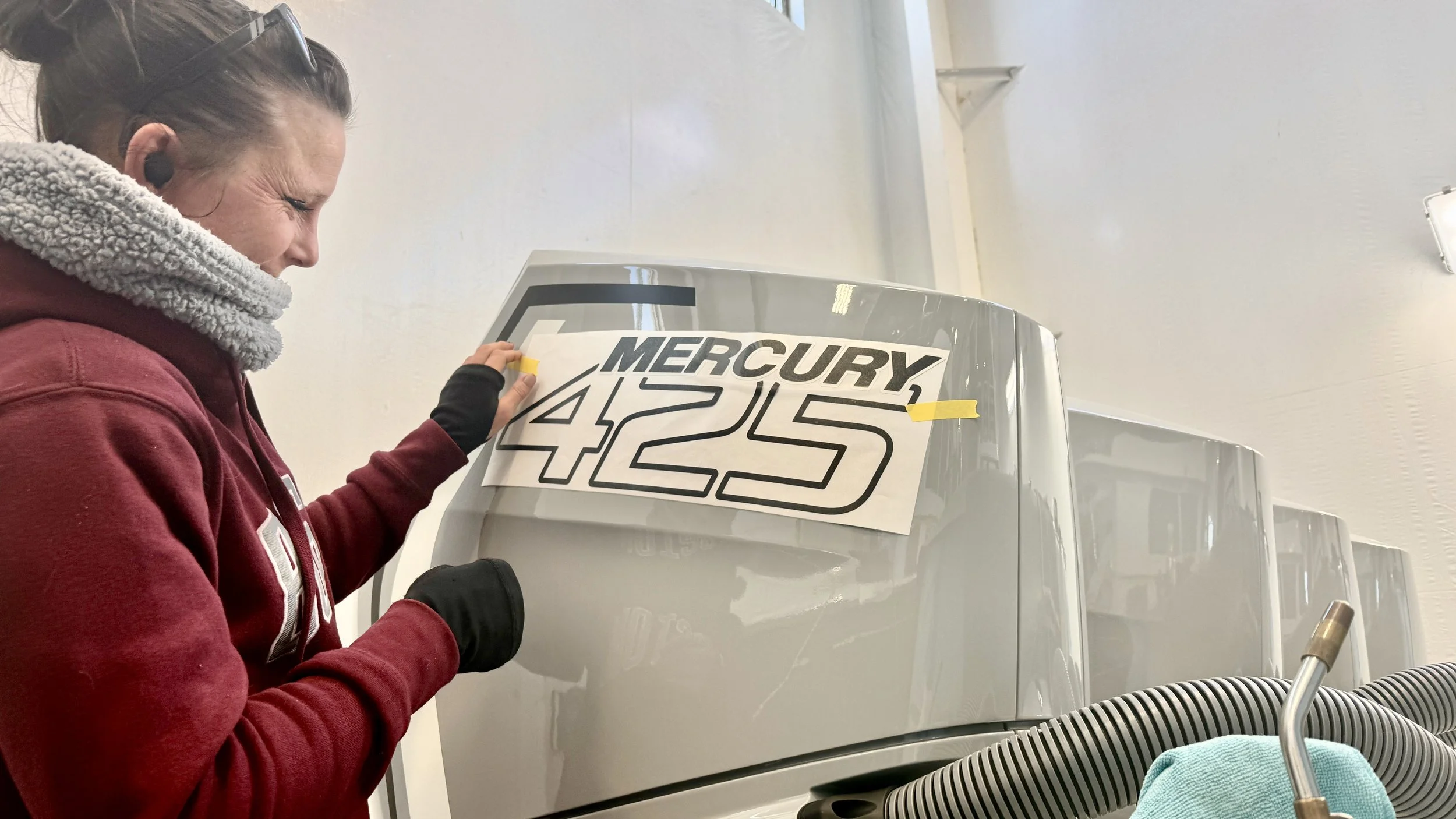 A woman with dark hair, wearing a red hoodie and a gray scarf, is applying a large decal with the words 'MERCURY 425' on a piece of equipment, possibly a racing boat or vehicle, inside a room with white walls.