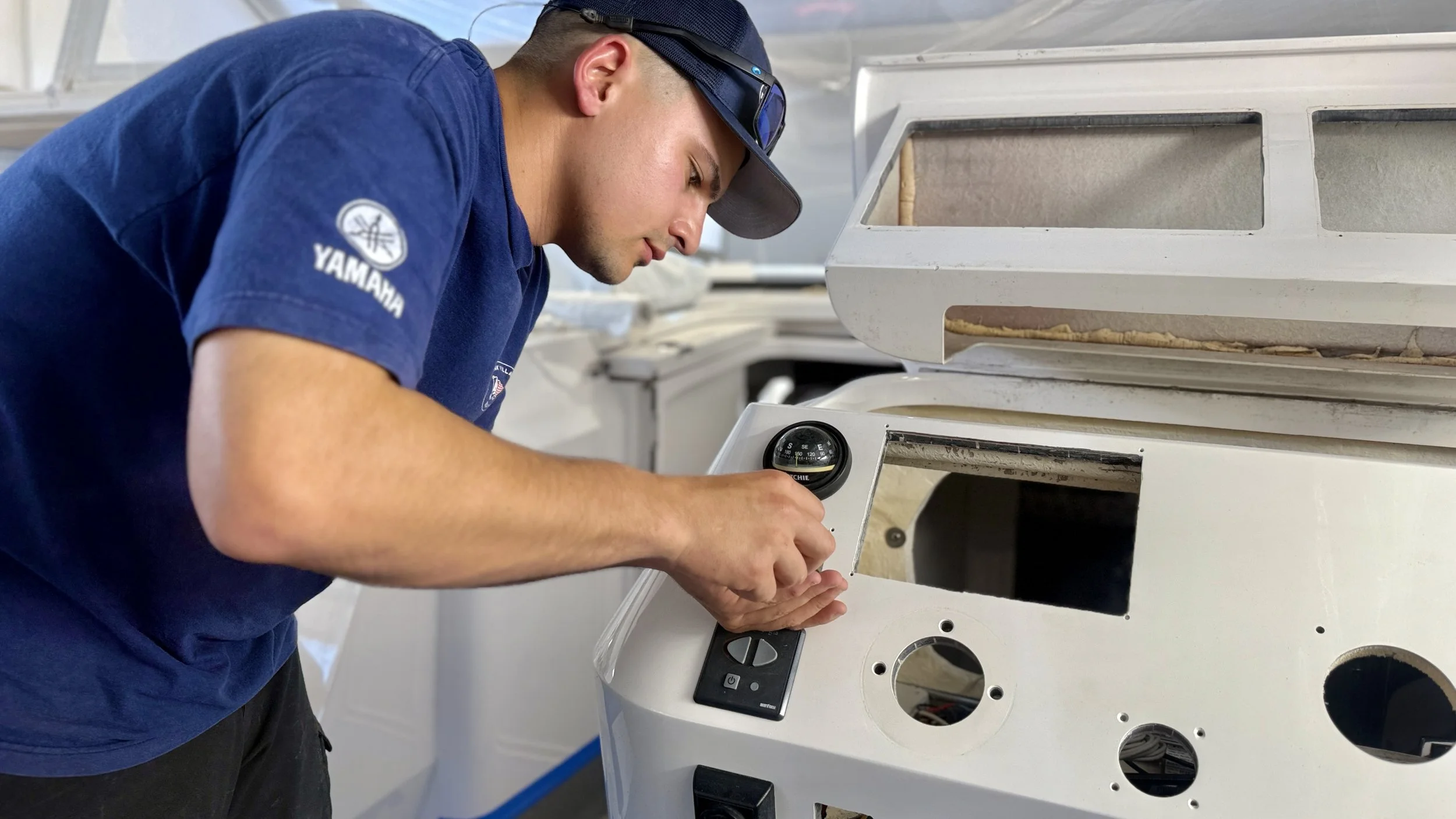 A technician wearing a Yamaha shirt and a cap inspects an electronic device with a gauge and digital controls inside a white ashy chassis, possibly working on a speaker or electronic equipment