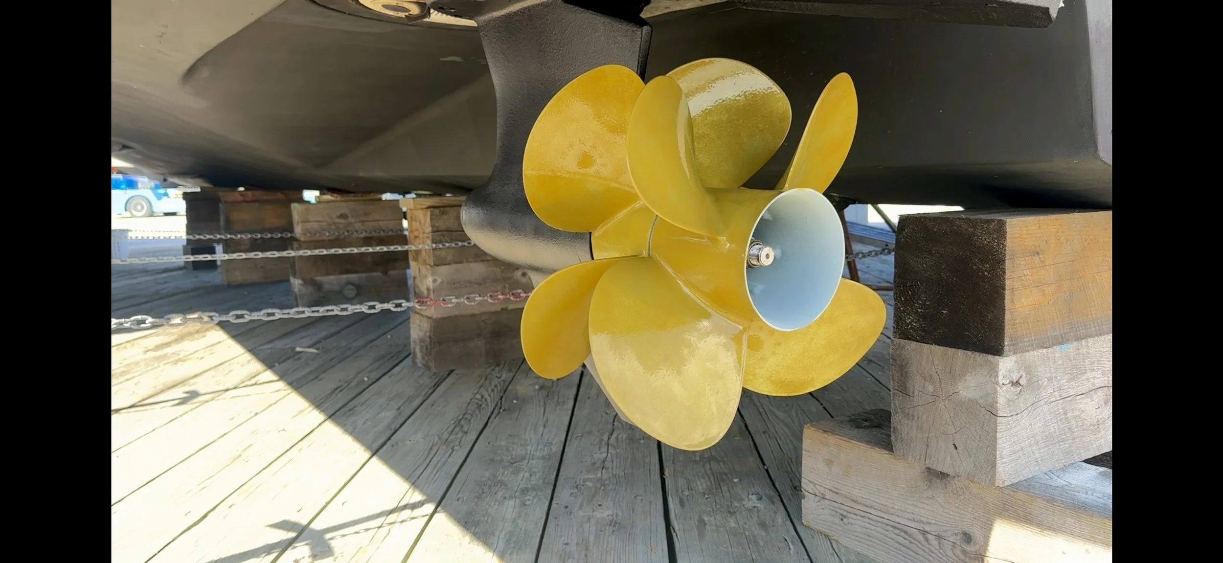 The image shows a boat propeller with yellow blades mounted on the boat's rudder, resting on wooden supports on a wooden dock. The background includes parts of the dock and some parked vehicles.