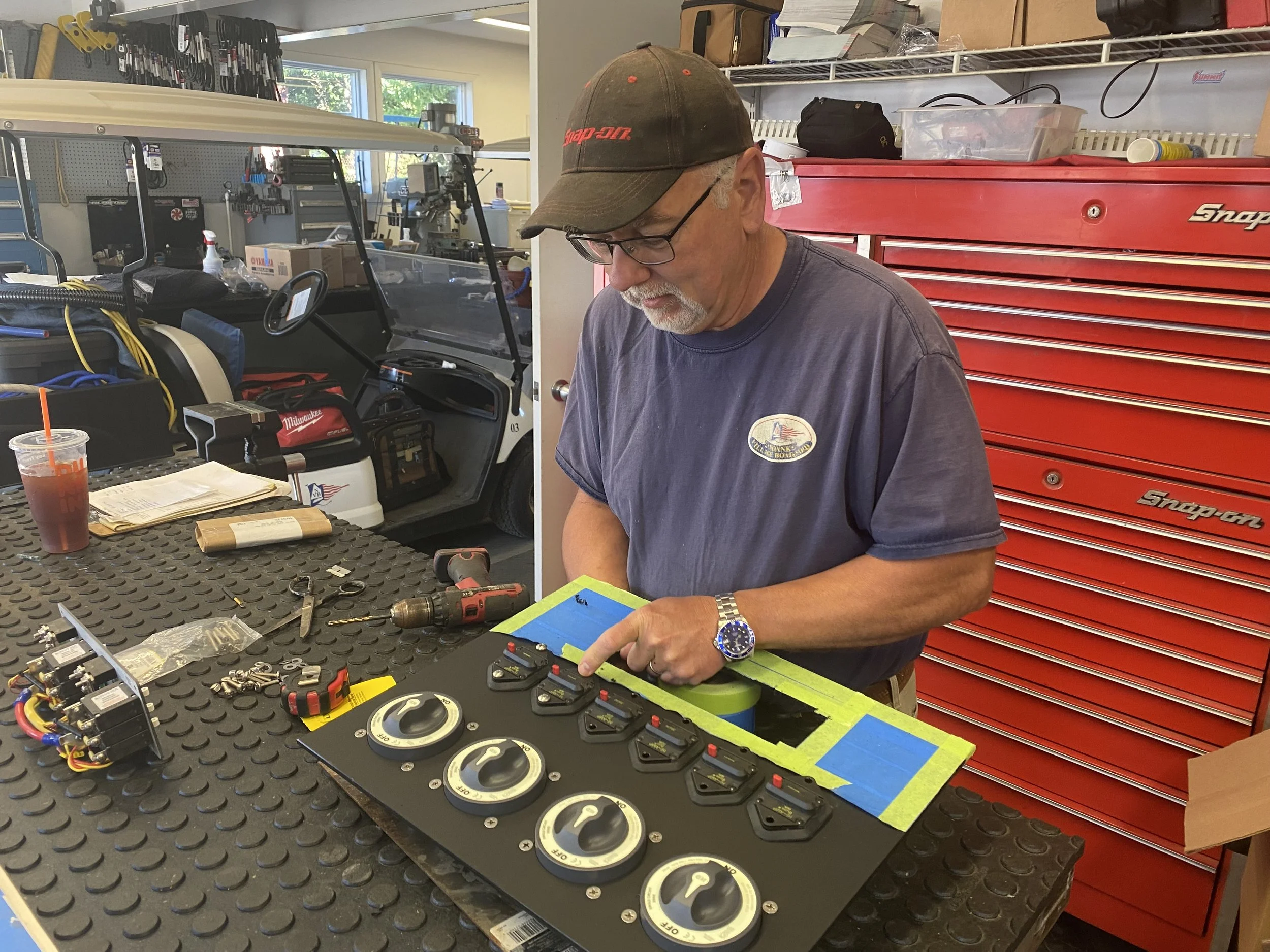 A man with glasses and a black cap working in a garage or workshop, surrounded by tools and equipment. He is standing at a workbench, possibly working on electrical or mechanical components, with various tools, wires, and parts spread out on the table. There is a large red tool chest behind him and a utility vehicle with equipment in the background.